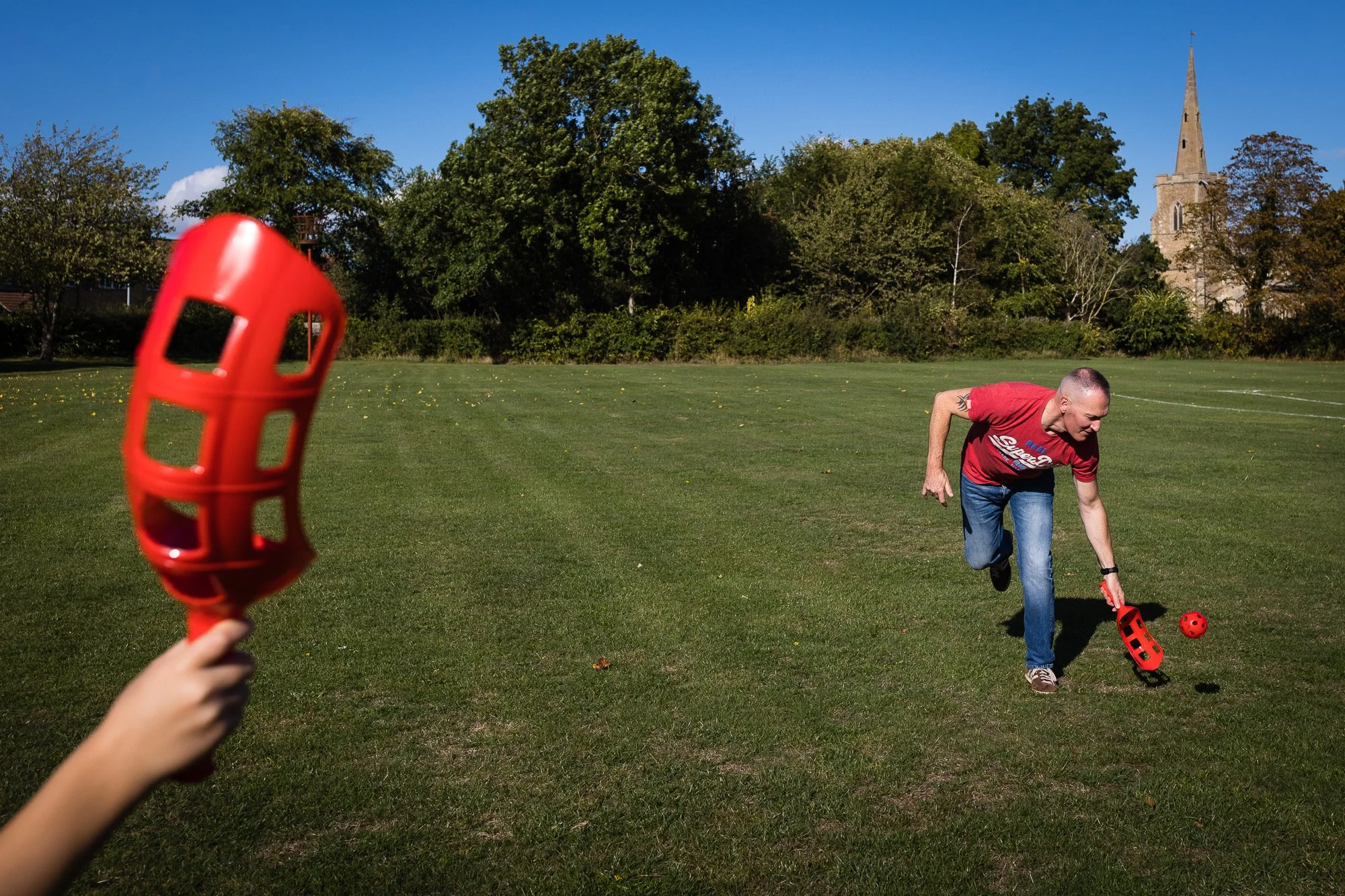A man in a red t-shirt and jeans playing croquet on a grassy field, with a hand holding a red croquet mallet in the foreground and a church steeple visible in the background.