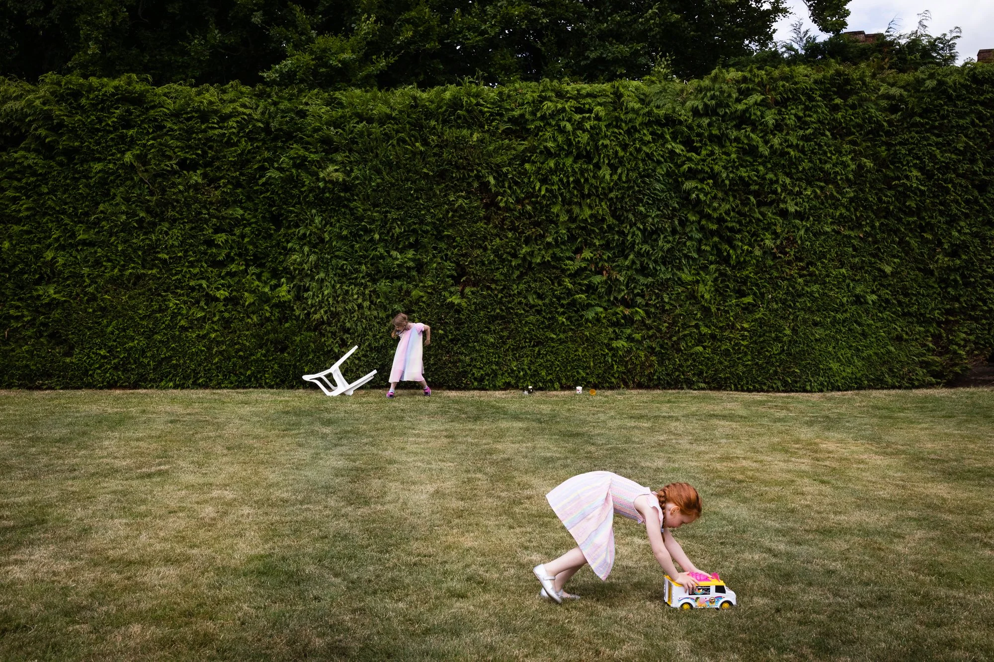 Two young girls playing on a lawn in front of a tall hedge. One girl with red hair is bending over a toy bus, while the other girl with blonde hair is in the background near a fallen white chair.