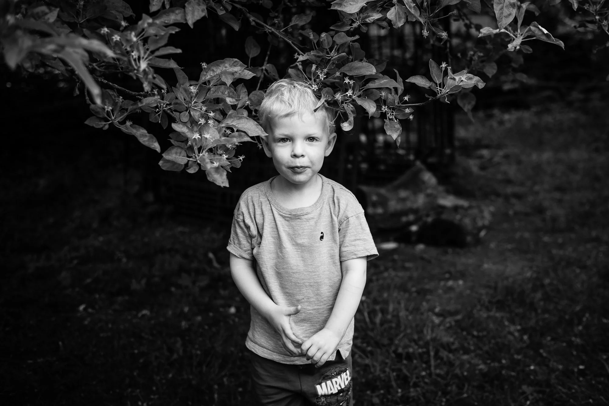 A young boy with light hair and a playful expression stands outdoors beneath tree branches, wearing a Marvel t-shirt.