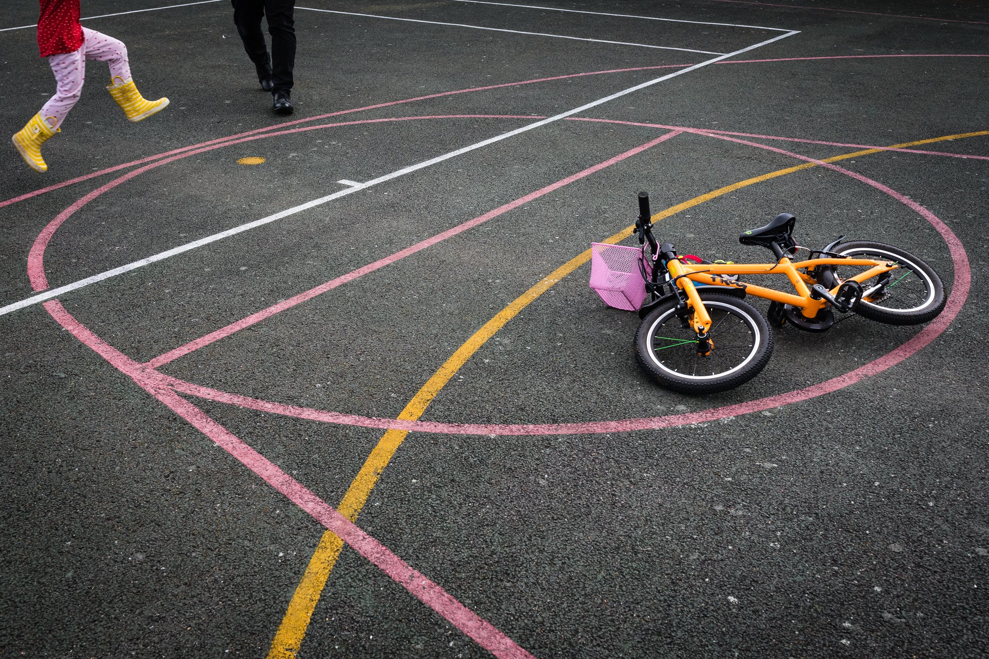A yellow and black children's bicycle with a pink basket on a marked basketball court, with a child and another person walking nearby.