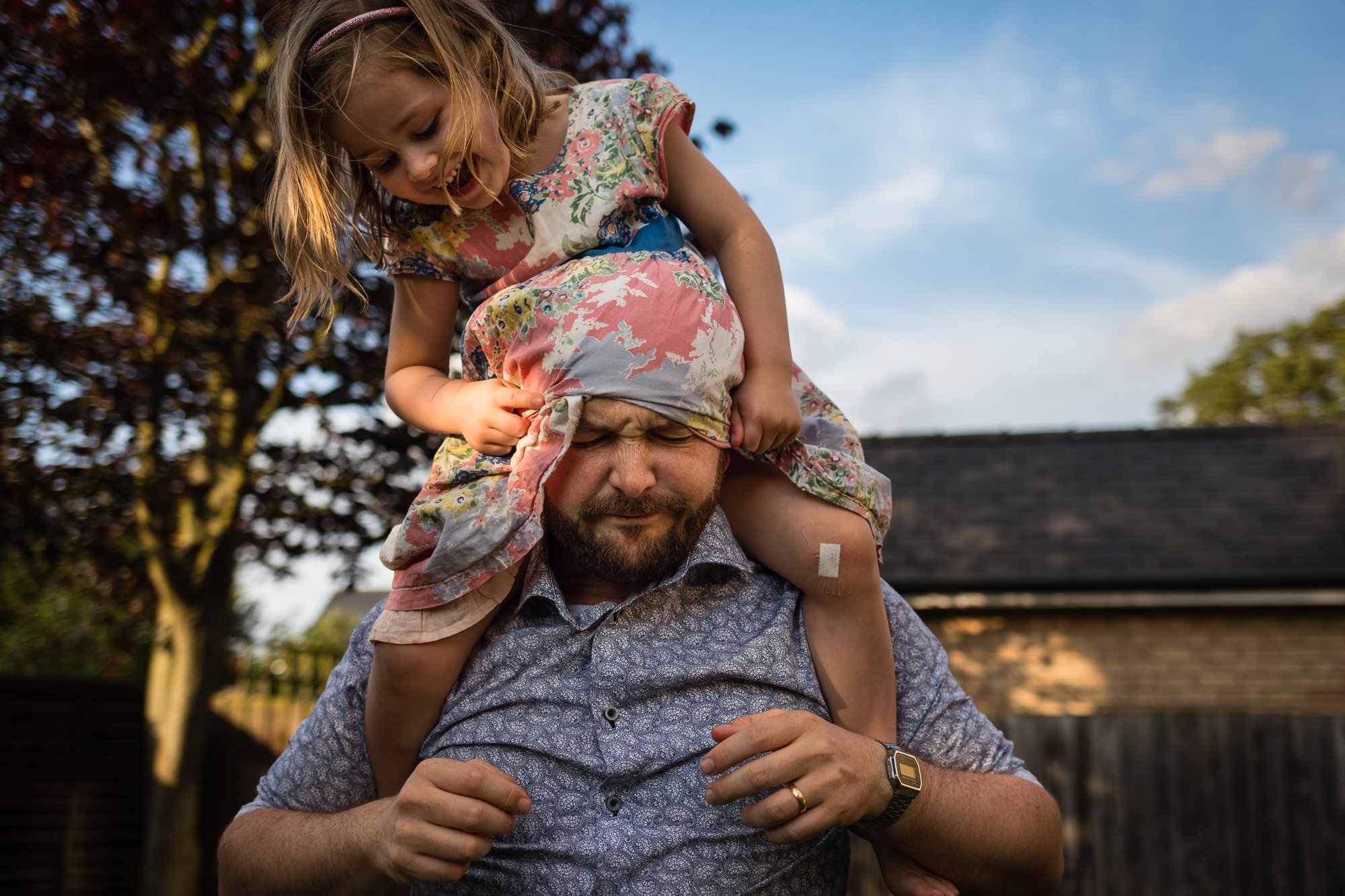 A young girl riding on her father's shoulders, pulling his bandana over his eyes during an outdoor activity in the backyard in the evening.
