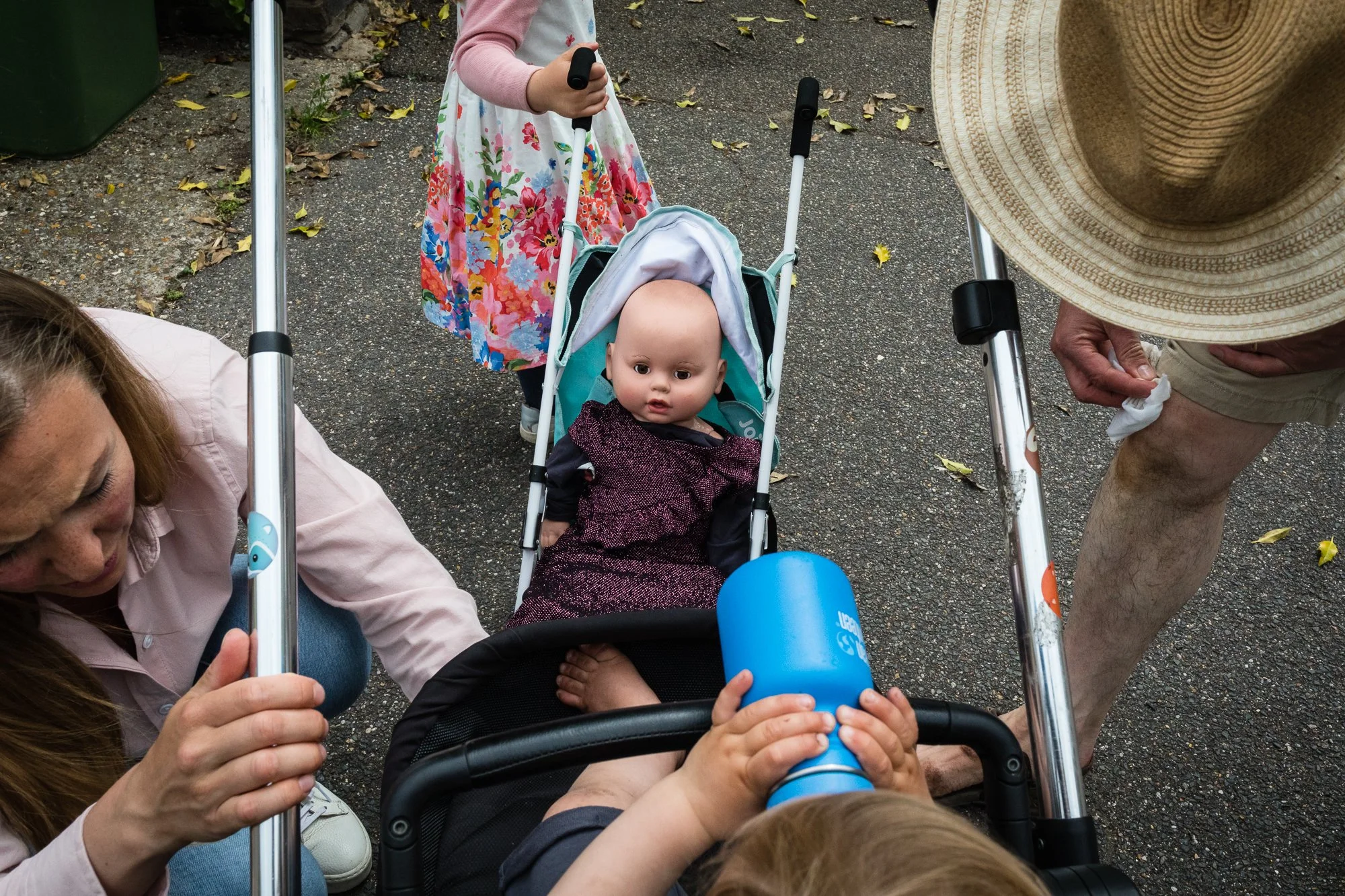 A young child in a stroller looking up at the camera, surrounded by adults and another child with a stroller, on an outdoor pavement with fallen leaves.
