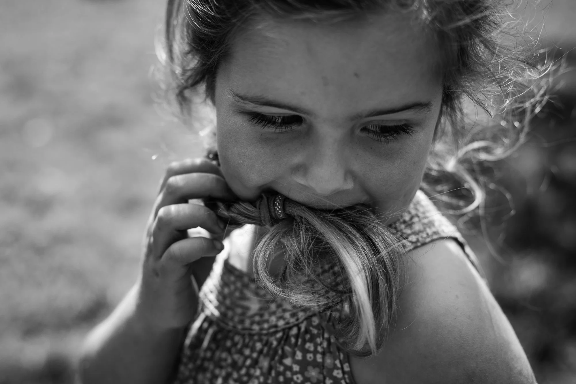A young girl with curly hair biting her own hair, holding it with one hand, outdoors on a sunny day.
