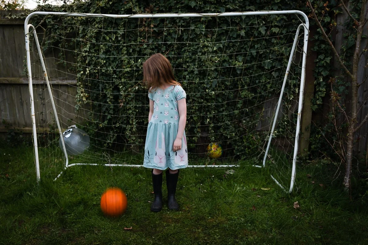 A young girl with long hair, wearing a light blue dress with bunny prints and black rain boots, stands in front of a small soccer goal on a grassy yard. She is looking down, with a soccer ball in motion near her. Another ball is visible behind the go