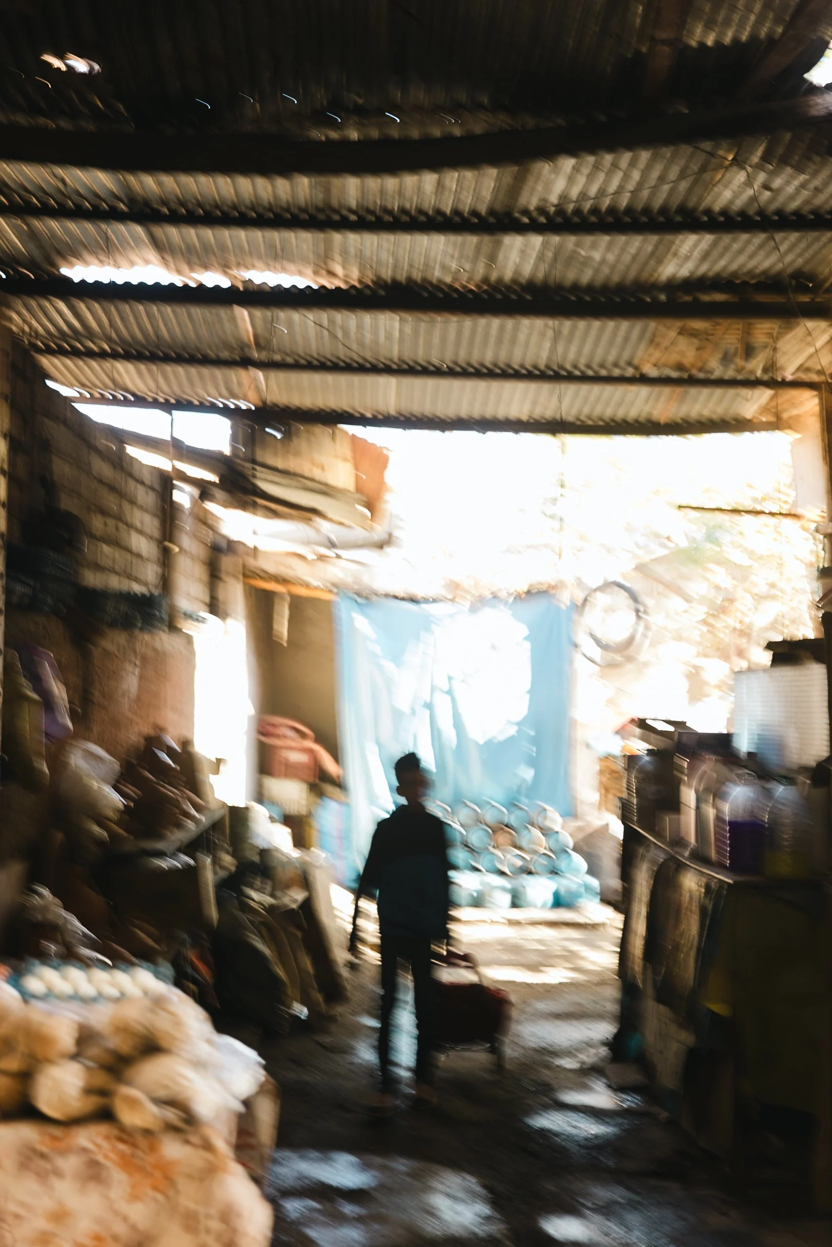 Inside a rustic shop or warehouse with a corrugated metal roof, a person stands with a red suitcase, silhouetted against sunlight, near shelves and stacks of goods. Blurred figures are seated on the left, and large blue sacks or covers hang in the ba