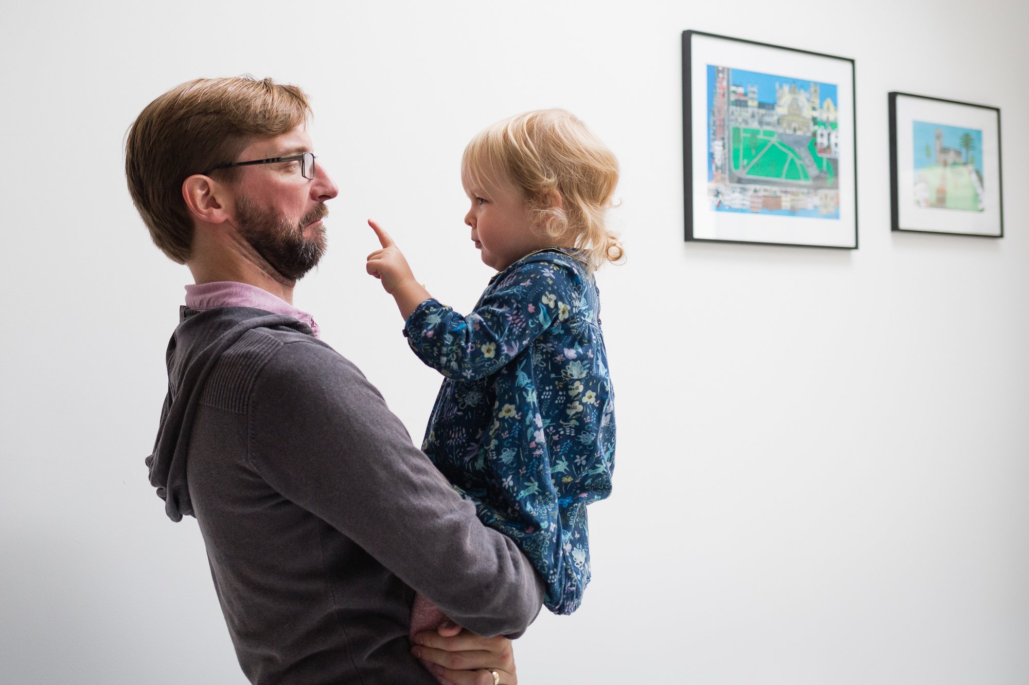 A man with glasses and a beard holding a young girl with blonde curly hair who is pointing her finger at his nose, standing in an art gallery with framed pictures on the wall.