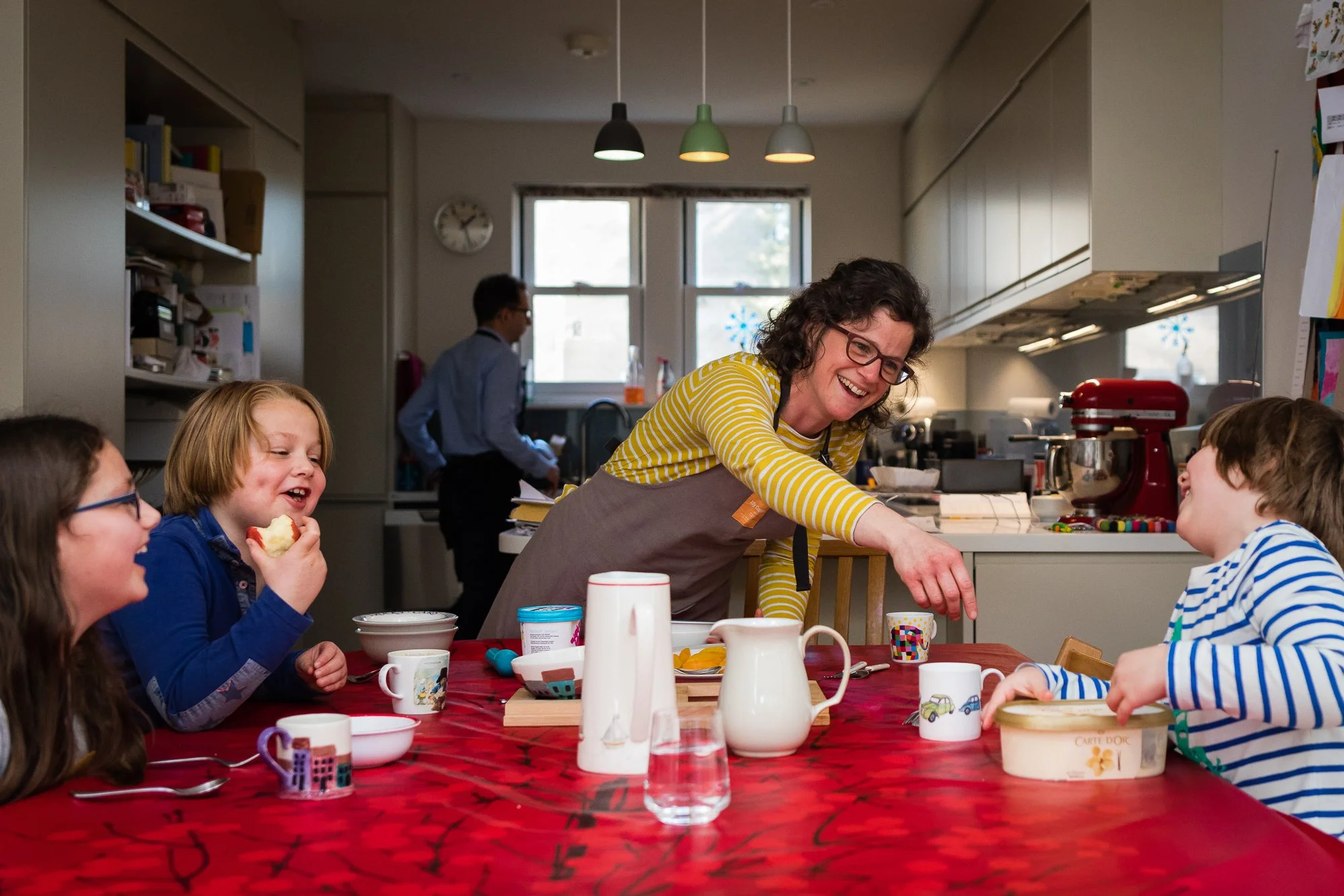 A woman serving breakfast to children at a table in a kitchen, with three children smiling and laughing. The table has mugs, bowls, and a pitcher, and the kitchen has a red stand mixer and other appliances.