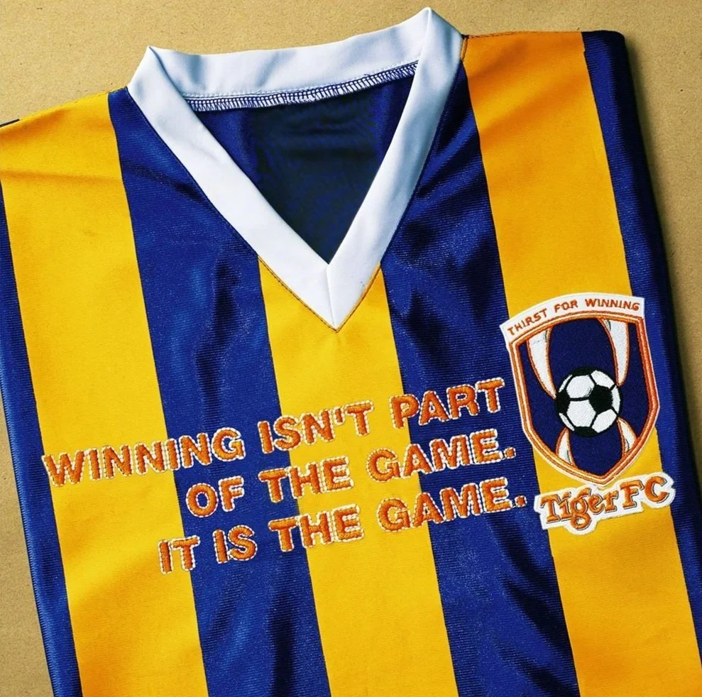 Close-up of a soccer jersey with blue and yellow vertical stripes, white collar, and embroidery that says "Winning isn't part of the game. It is the game." along with a crest featuring a soccer ball and a tiger's face, and text "Tiger FC".