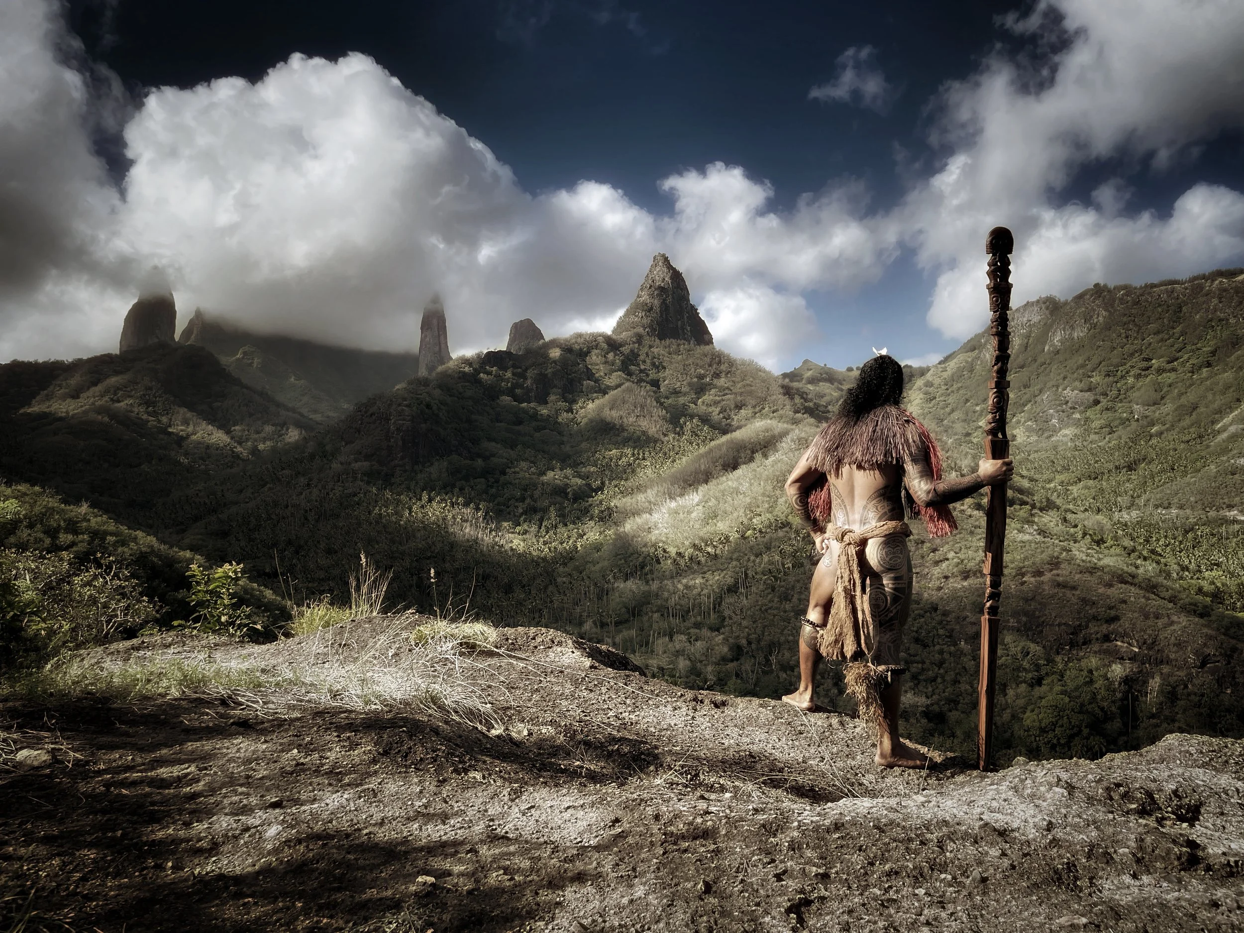 Une personne portant des vêtements traditionnels et tenant un bâton regarde un paysage de montagnes verdoyantes sous un ciel partiellement nuageux.