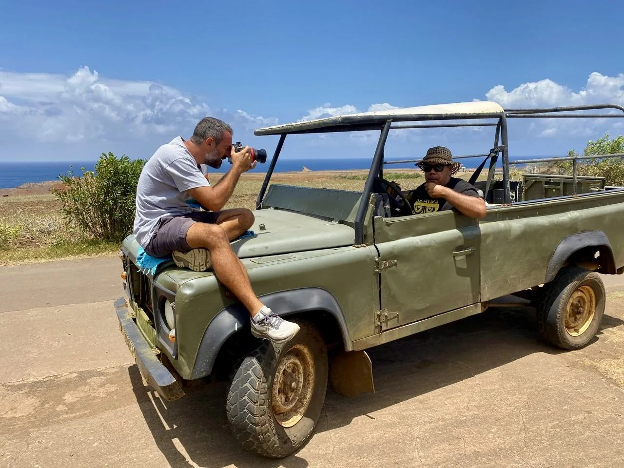 Deux hommes avec une Jeep ancienne sur une route côtière, l'un prenant des photos, l'autre conduisant. Paysage océanique en arrière-plan.