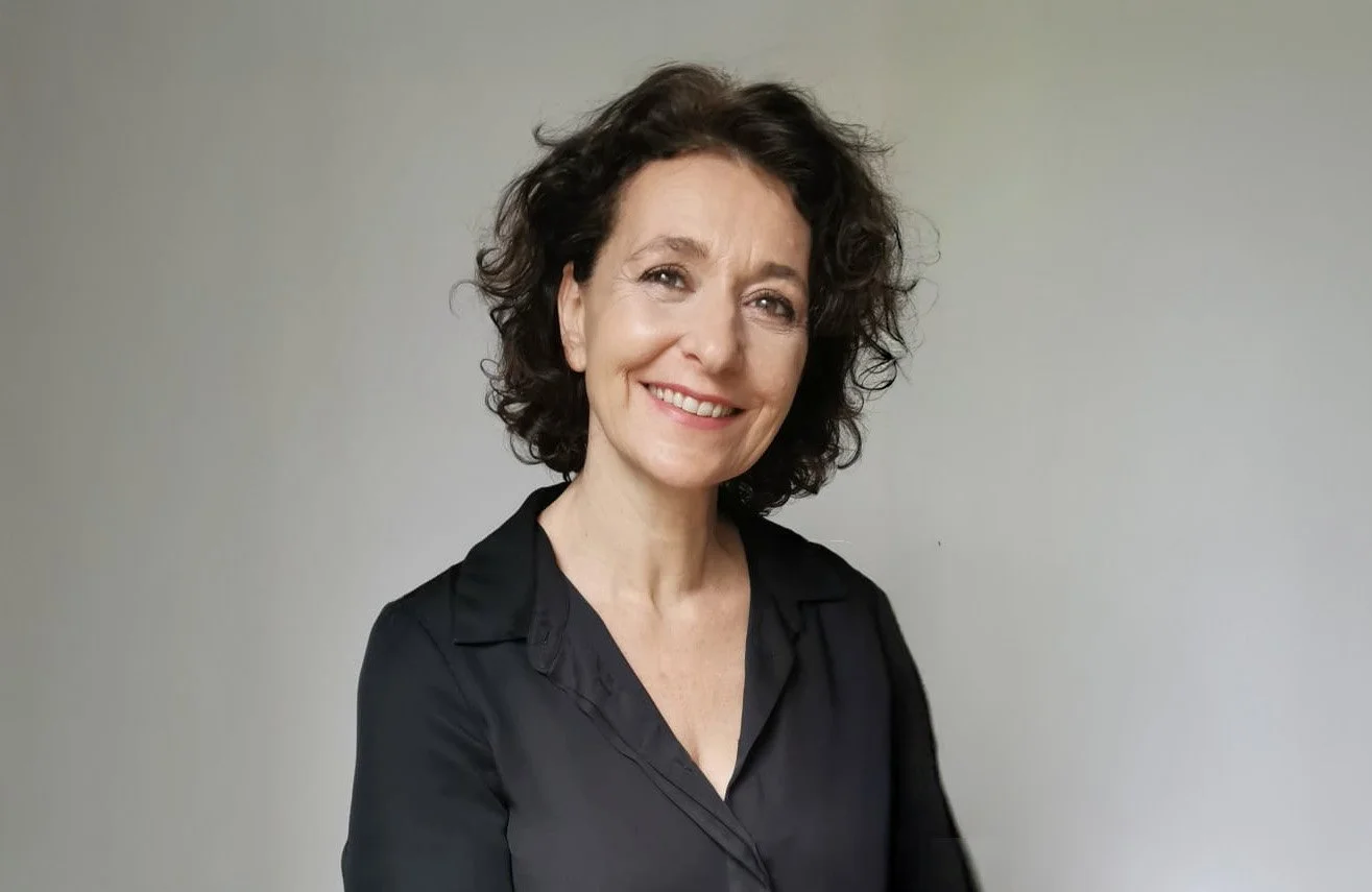 Femme souriante aux cheveux bouclés dans un chemisier noir sur fond neutre.