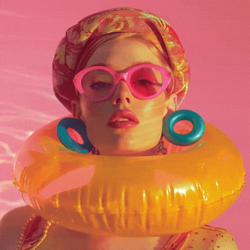 A woman with blonde hair wearing pink sunglasses, large turquoise earrings, and a colorful headscarf, surrounded by a yellow inflatable pool ring on a pink background.