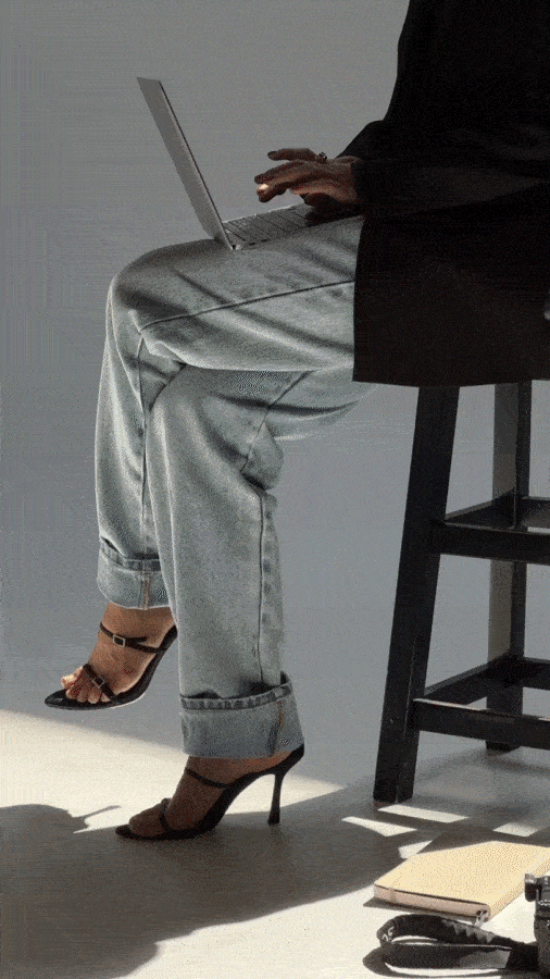 Person sitting on a chair, working on a laptop, wearing high heels and loose jeans, with a desk and miscellaneous office items nearby.