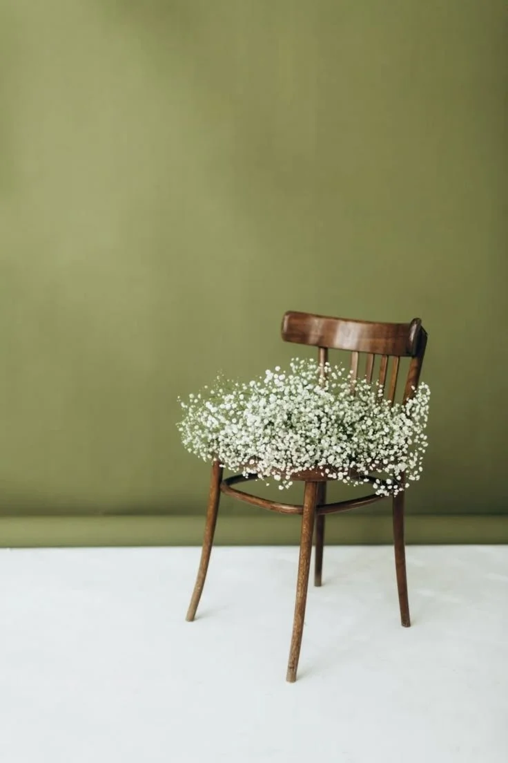 A wooden chair with a bouquet of small white flowers resting on its seat, set against a plain green background.
