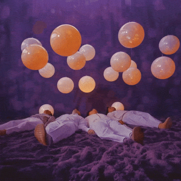 Four people lying on a purple ground in white clothing beneath white and orange balloons suspended in the air.