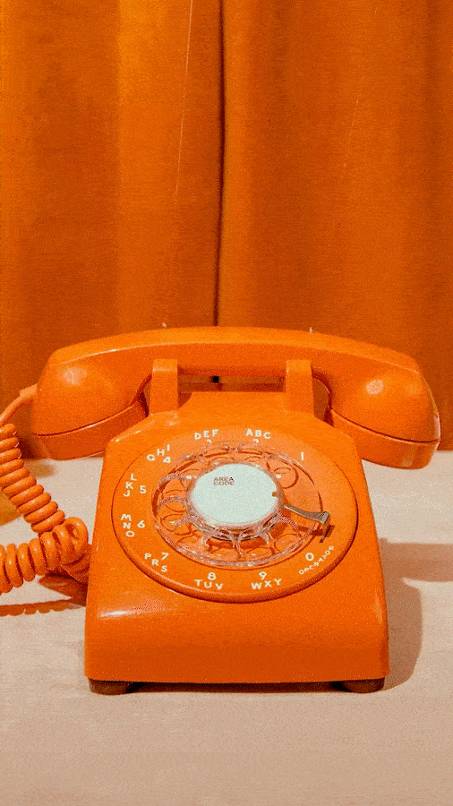 An orange rotary telephone with a coiled cord, set against a warm-colored curtain background.