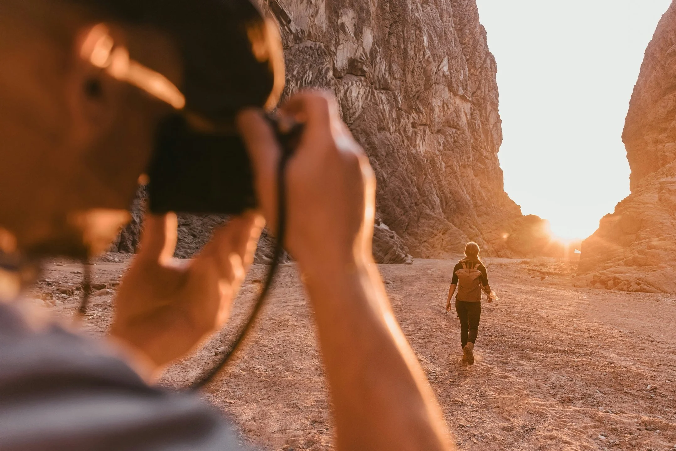 Person taking a photo of another person walking through a canyon during sunset.