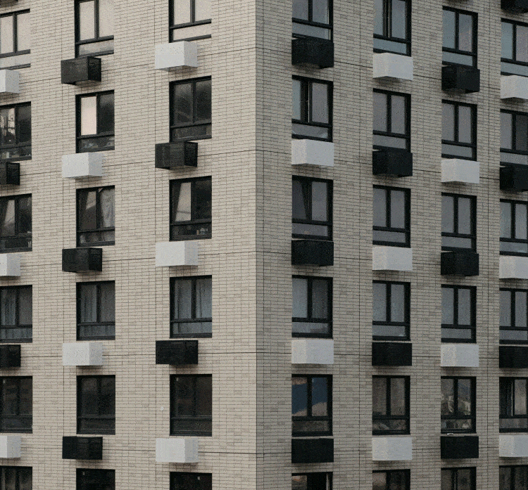 Close-up of a multi-story building facade with beige brick exterior, black-framed windows, and small black and white protruding window boxes.