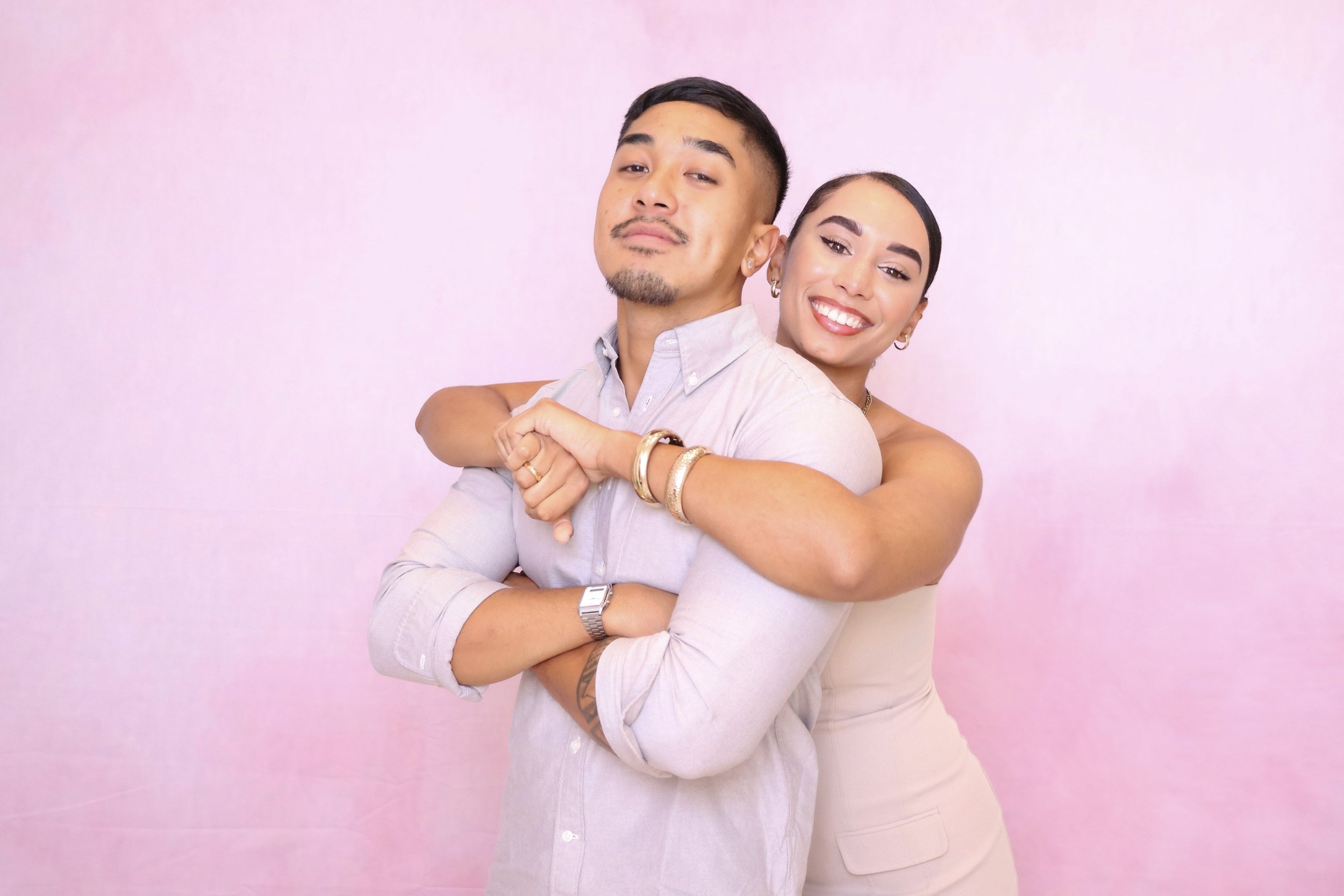 A woman with dark hair and a man with short dark hair and facial hair pose together against a light pink background. The woman is smiling while hugging the man from behind. The man has arms crossed, wearing a light-colored button-up shirt and has a confident expression.