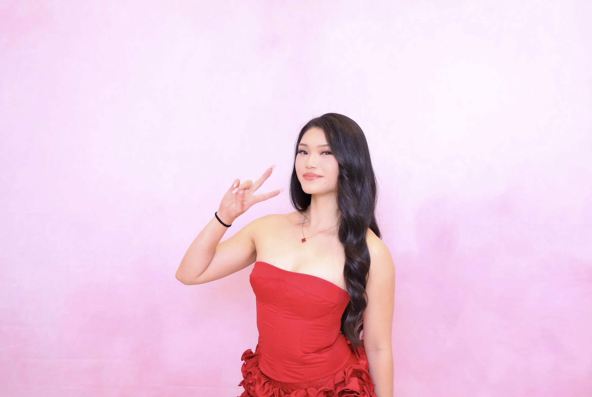 Young woman in a red strapless dress posing against a pink background, making a peace sign with her right hand.