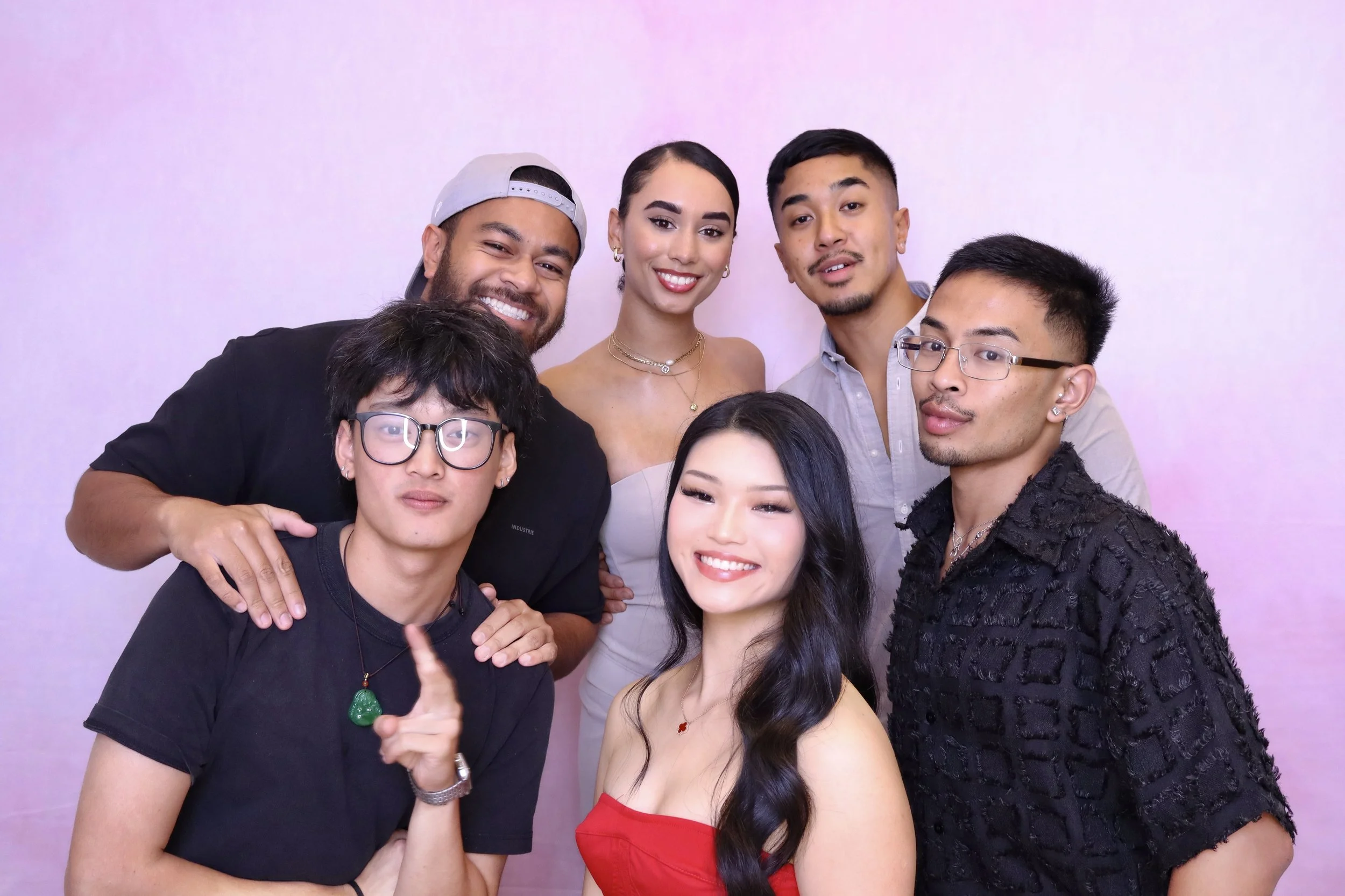 Group of seven diverse young adults smiling and posing together in front of a plain light pink background, dressed in casual and semi-formal clothing.