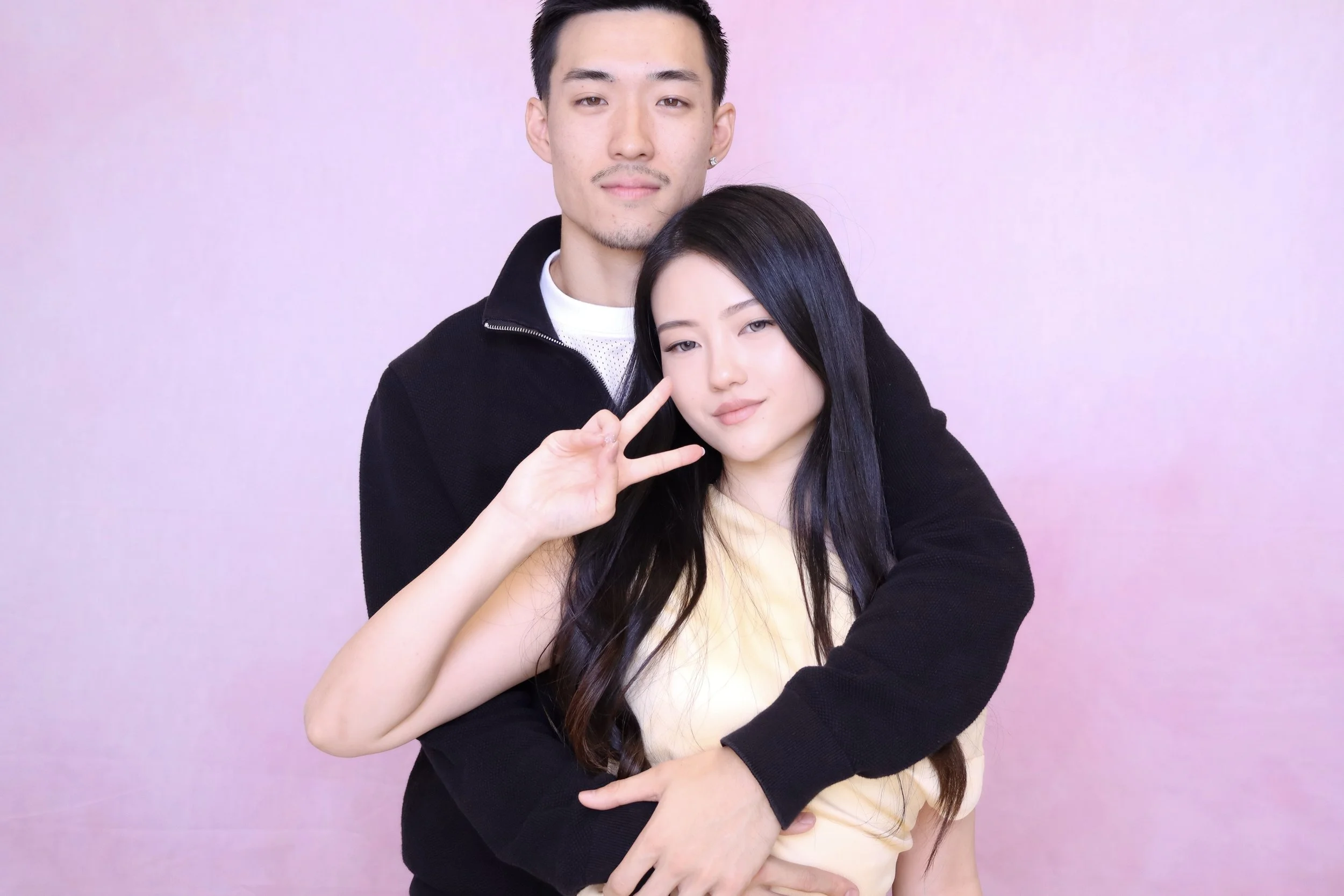 A young man and woman posing together against a pink background. The man is standing behind and hugging the woman, who is making a peace sign with her right hand near her face.