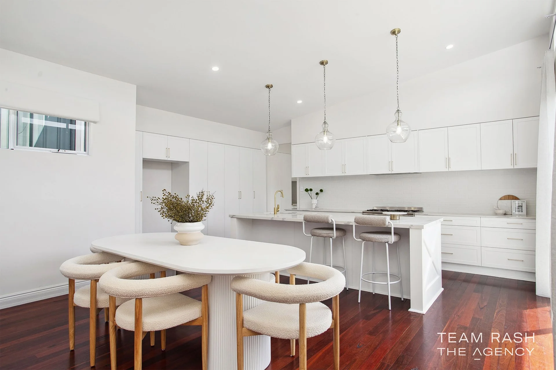 Modern, minimalist kitchen and dining area with white cabinets, white walls, and a white oval table with six upholstered chairs on a dark hardwood floor. Three pendant lights hang above the kitchen island, which has a gold faucet and beige barstools.