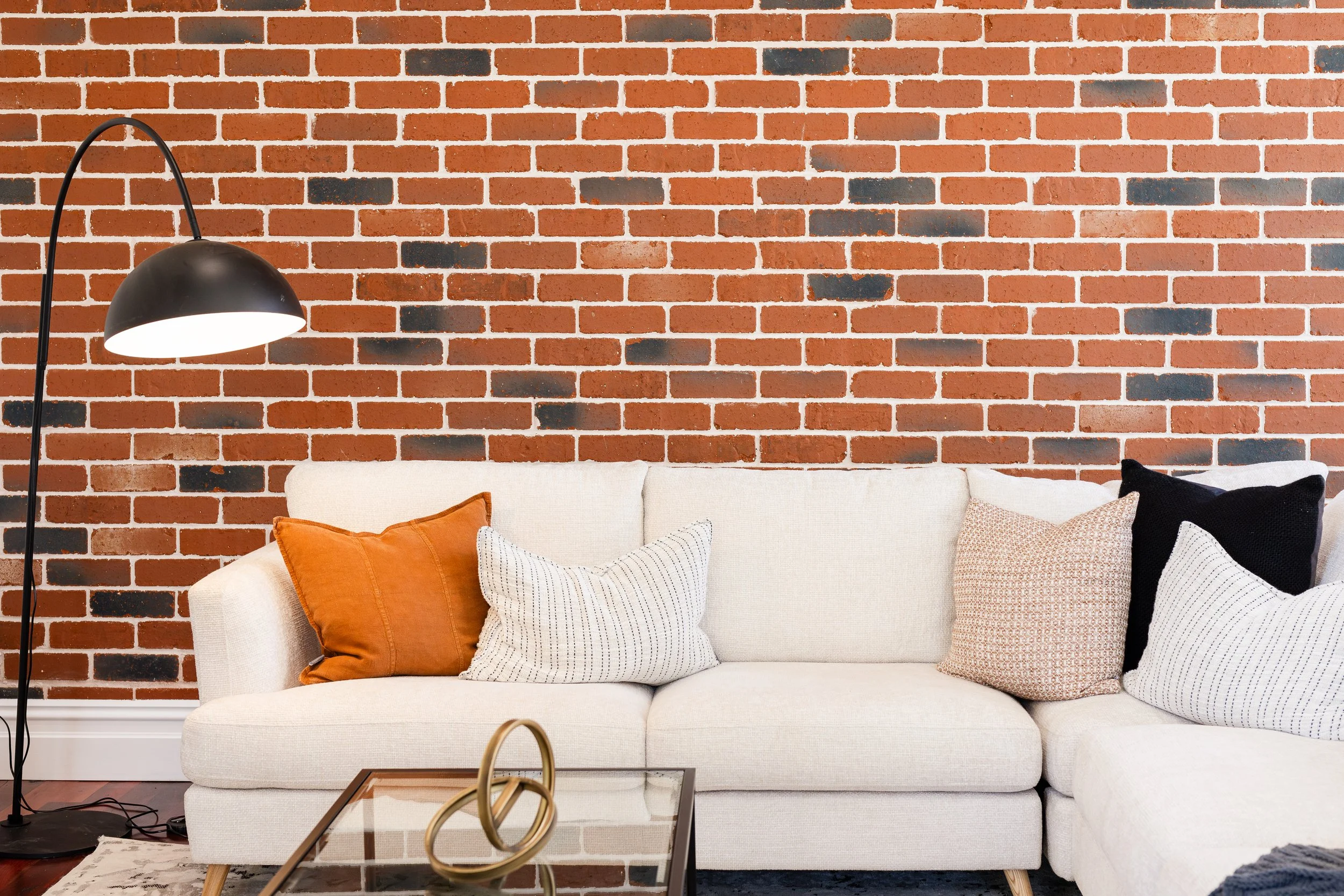 Modern living room with a cream sofa decorated with various pillows, a glass coffee table with a decorative gold sculpture, a black floor lamp, and an exposed brick wall in the background.