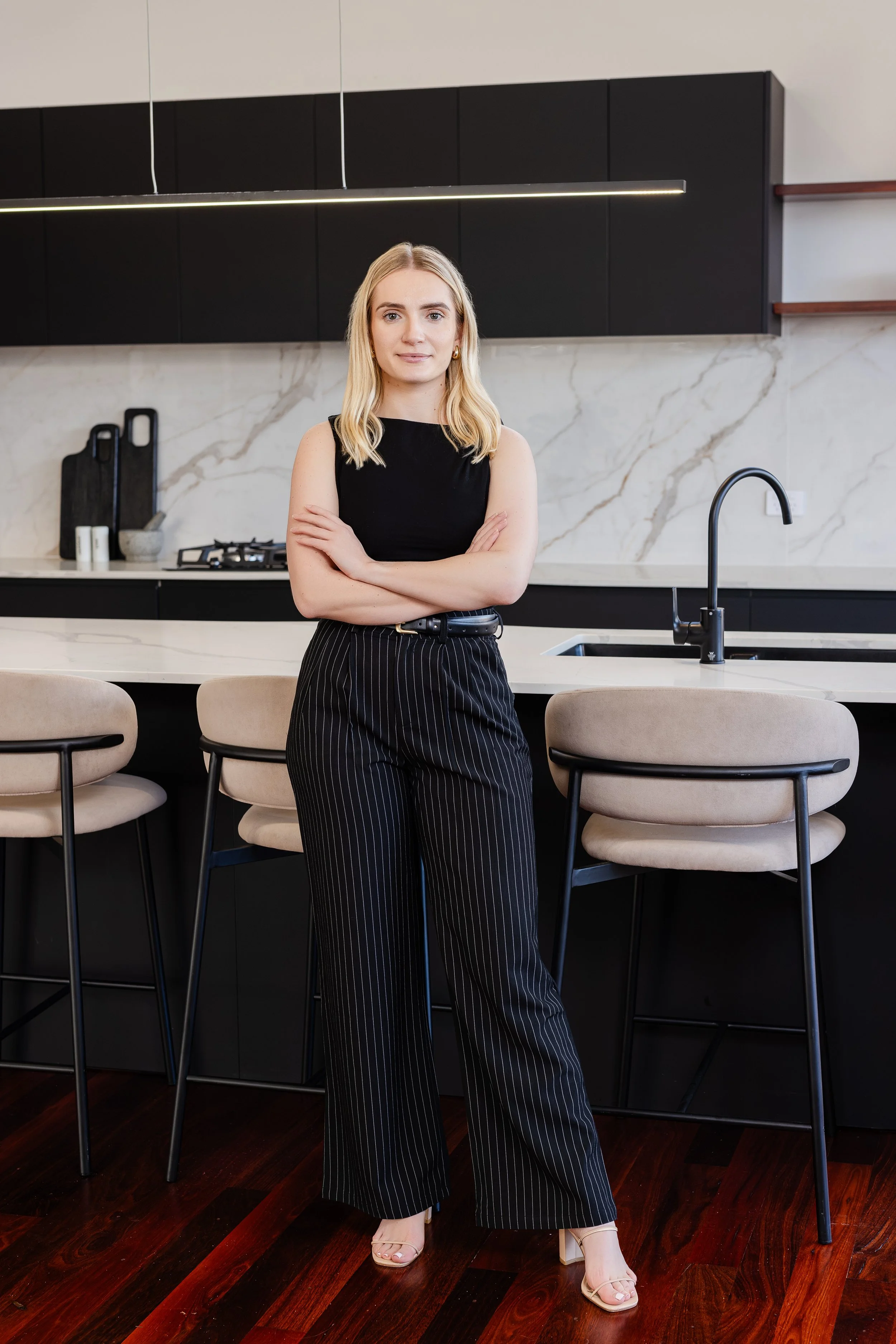 A young woman standing in a modern kitchen with her arms crossed, dressed in black sleeveless top and black pinstripe pants.