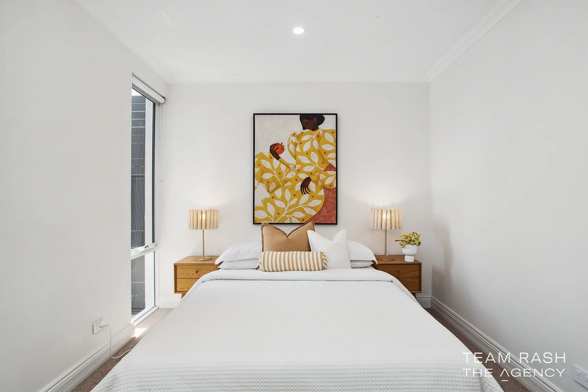 A neatly made bedroom with a white bed, beige and white pillows, wooden nightstands on each side, matching lamps, a window on the left, and a colorful abstract art piece above the bed.