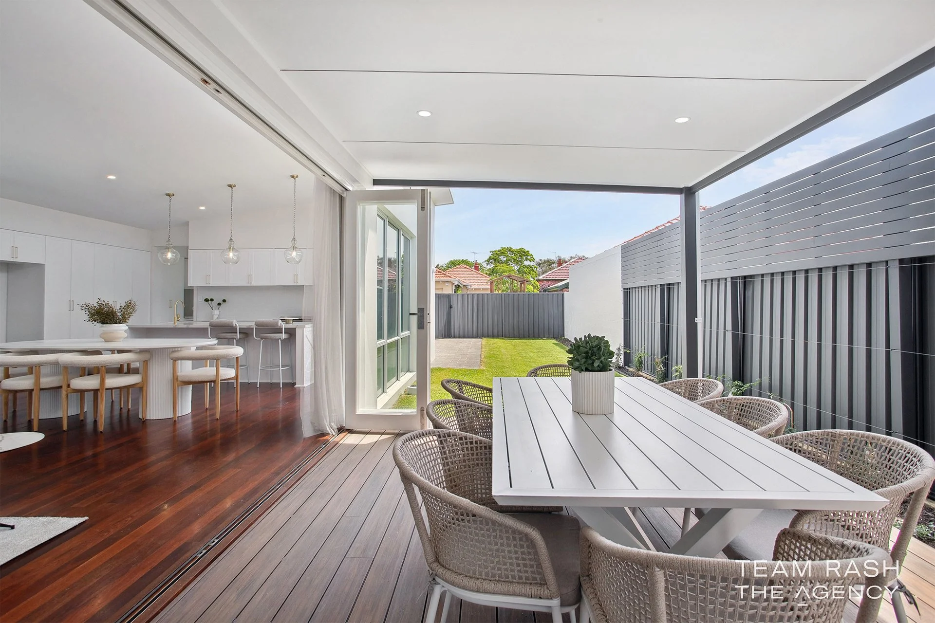 Indoor-outdoor living space with a wooden deck, white dining table with woven chairs, and a view of a backyard with a lawn and gray fencing