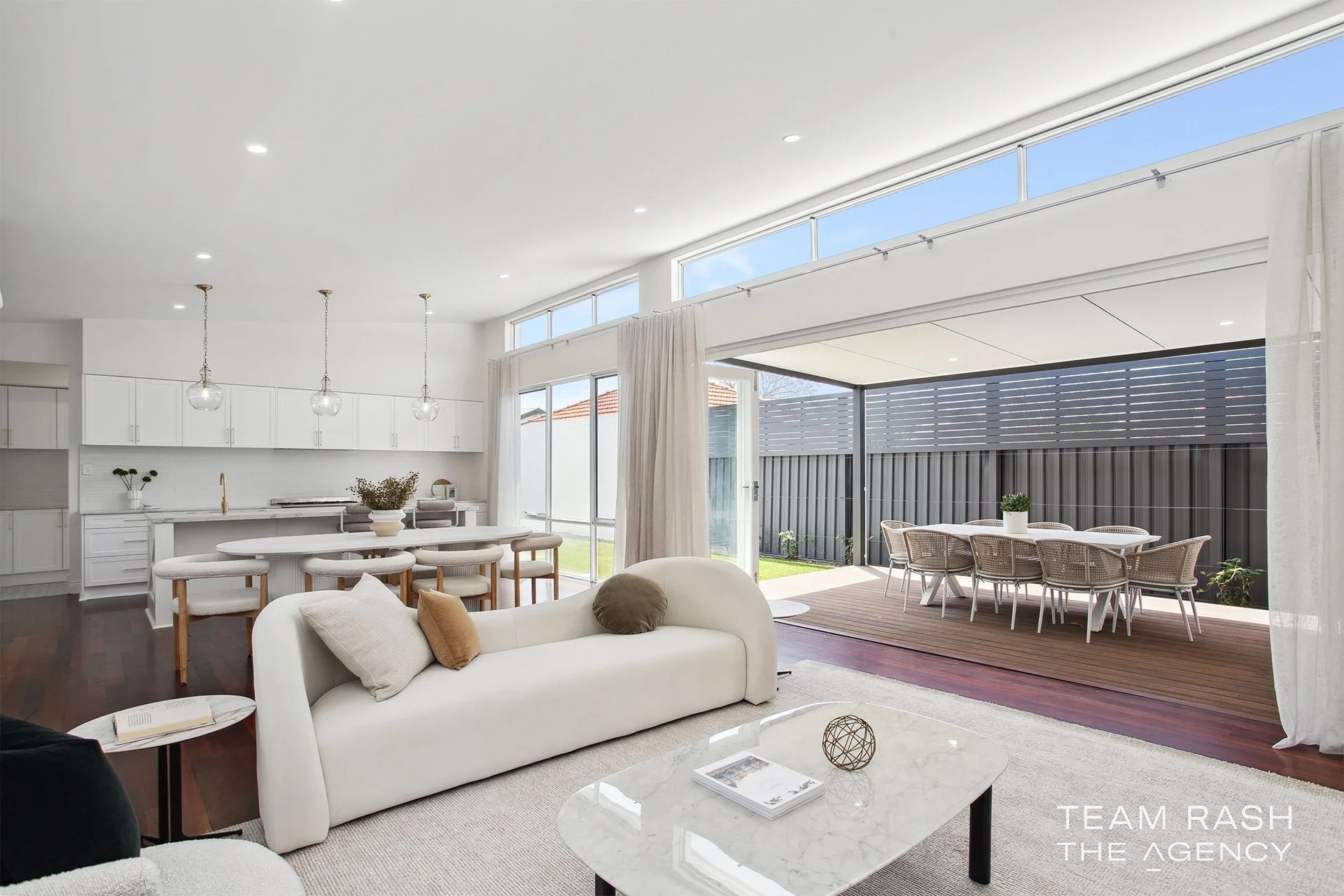 Modern open-concept living room and kitchen with sliding glass doors leading to outdoor patio with dining table and chairs, white sofa with pillows, and minimalistic decor.