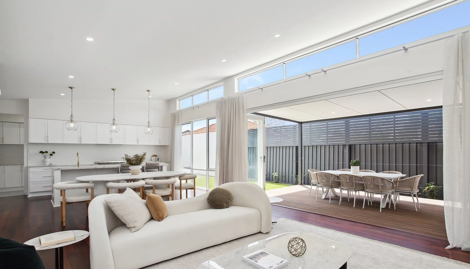 Modern open-plan living space with white kitchen, dining area, and outdoor patio with wicker table and chairs.