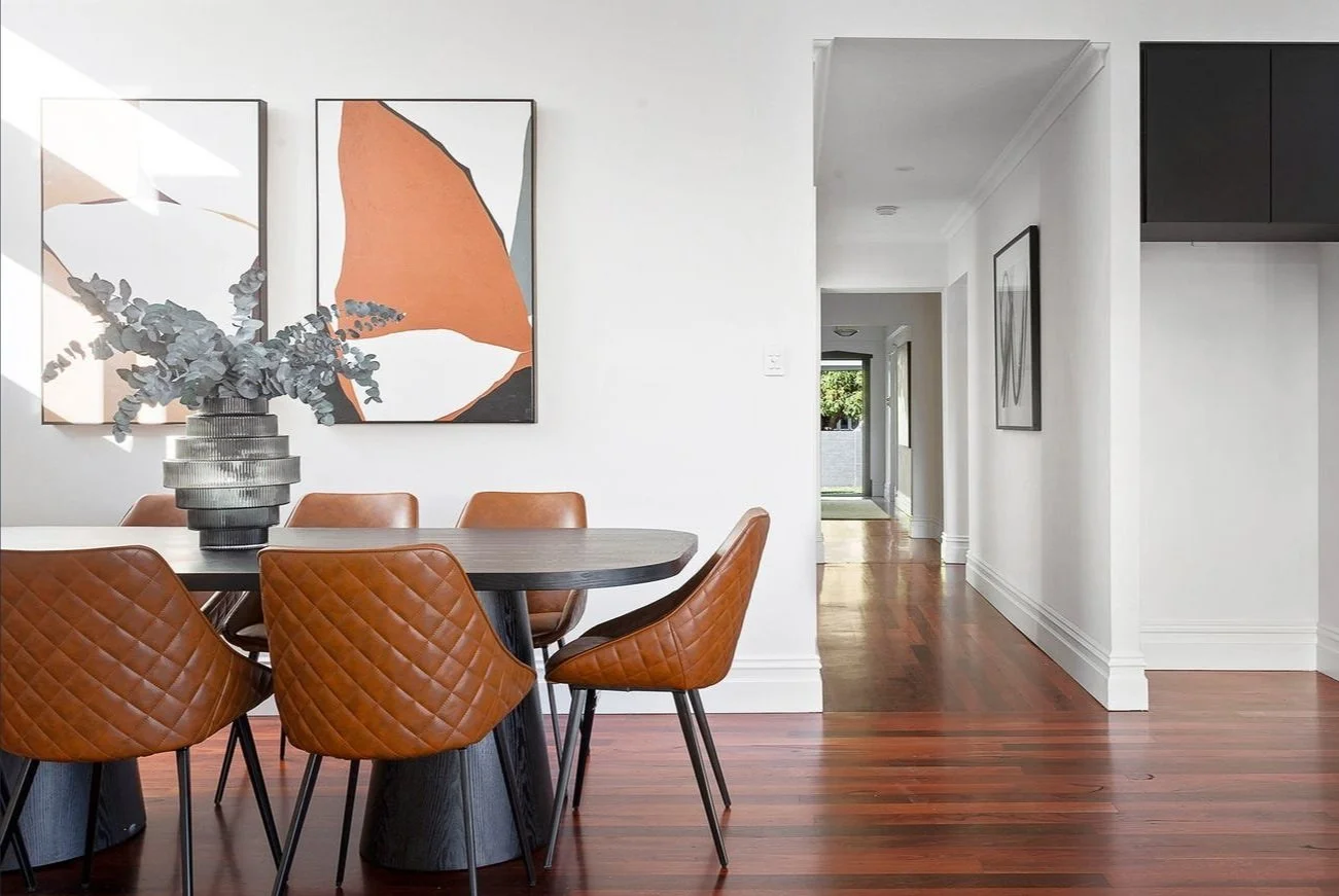 A dining area with a black oval table, six brown quilted leather chairs, a vase with gray eucalyptus, and abstract artwork on the white wall. A hallway with wooden floor extends into the background.