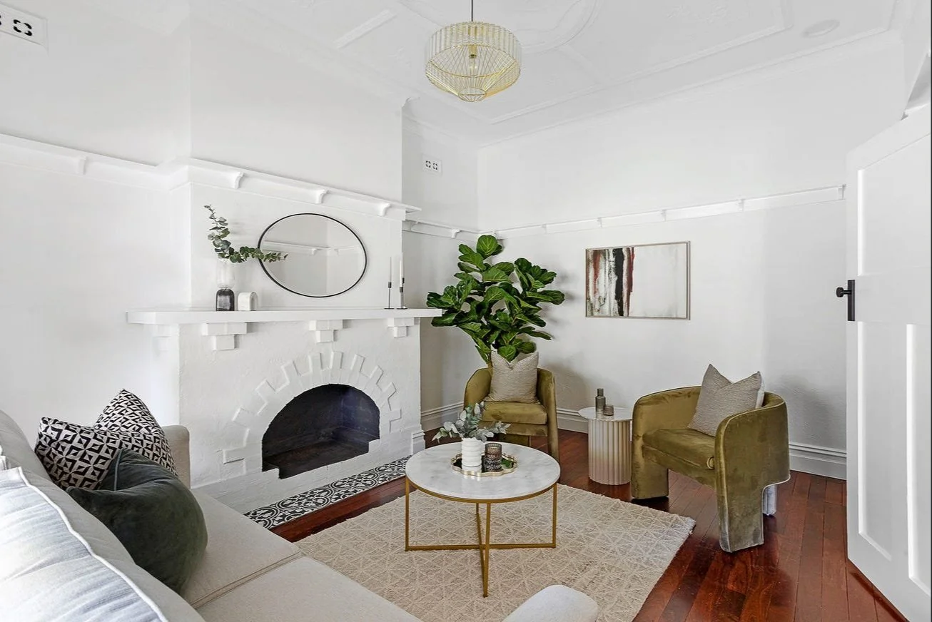 Living room with white walls, a fireplace with a white mantel and black opening, a large green potted plant, two olive green velvet armchairs, a beige sofa with patterned pillows, a round marble-top coffee table, and wall art.