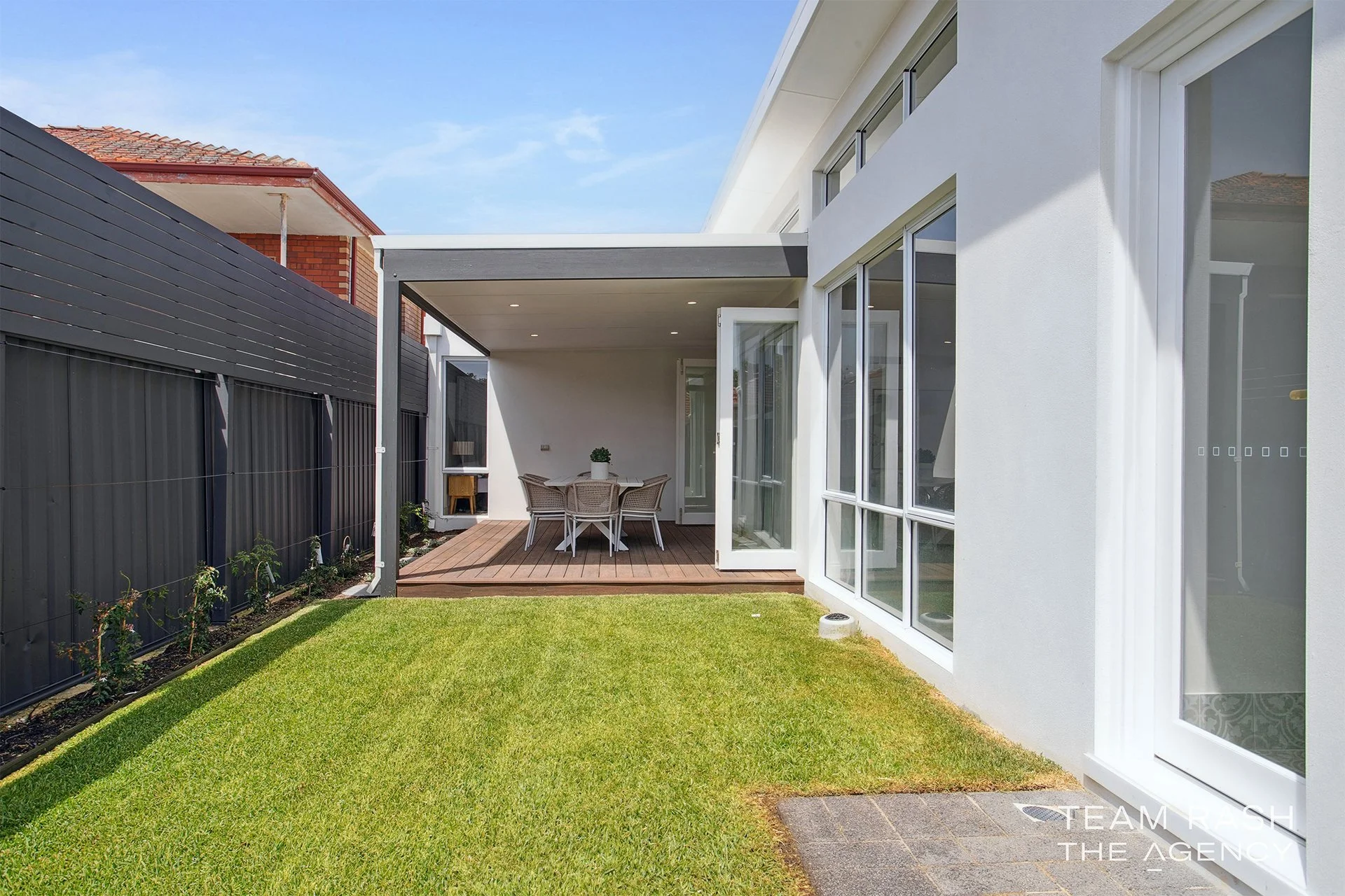 Modern backyard with a green lawn, a wooden patio with outdoor furniture, a black fence, and a white house with large windows and a covered porch.
