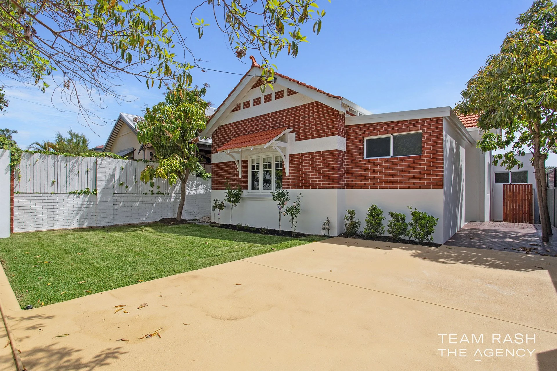 A modern house with a brick and white exterior, a small front yard with green grass and trees, a concrete driveway, and a white fence under a clear blue sky.