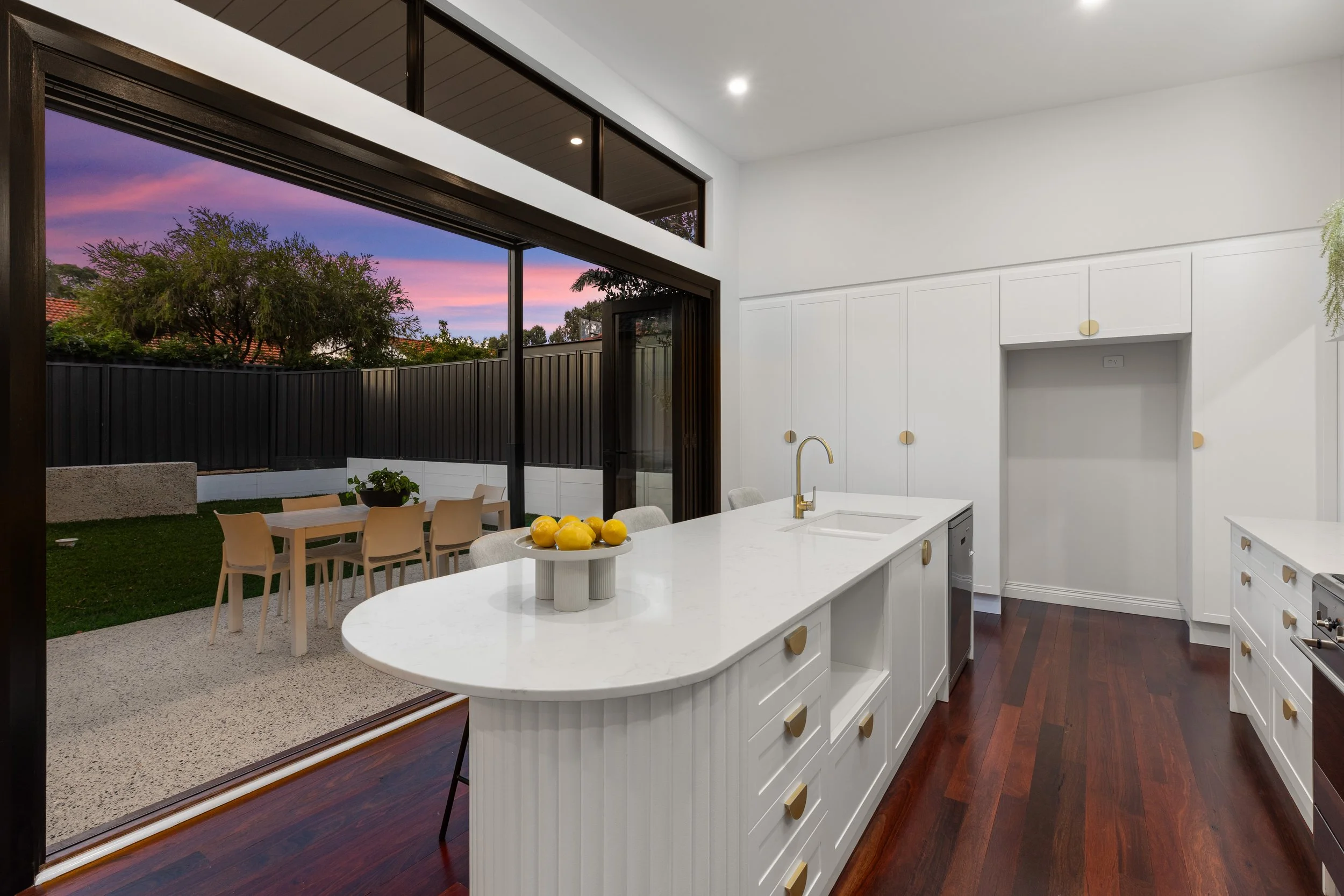 Modern kitchen with white cabinets, marble island with a sink, and a view of the backyard patio with dining table and chairs at sunset.