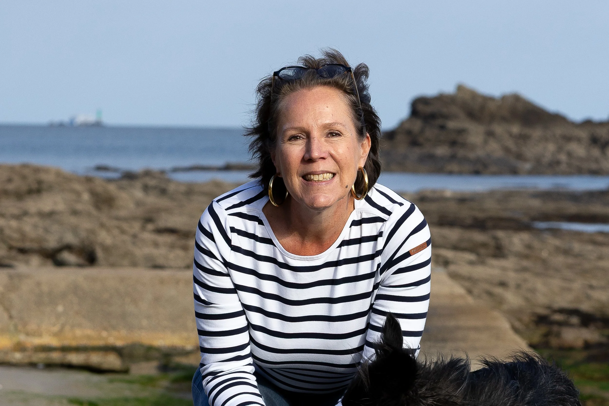Femme souriante portant un t-shirt à rayures noires et blanches, avec des lunettes sur la tête, à la plage avec des rochers et la mer en arrière-plan.
