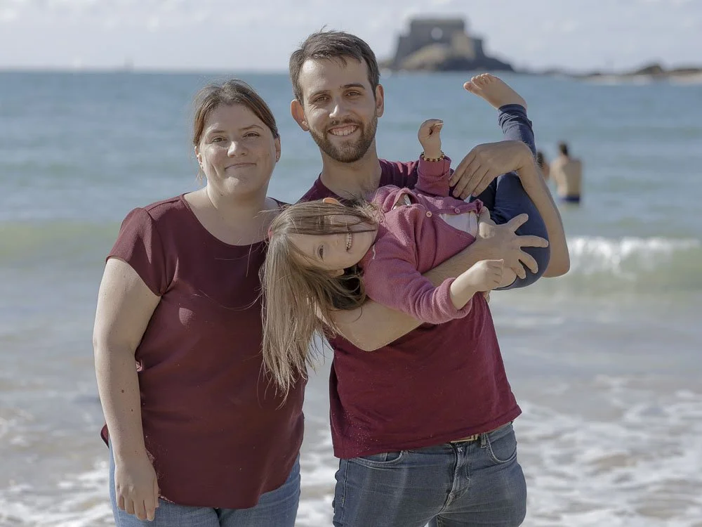 Famille au bord de la mer avec un homme, une femme et une fille portant des vêtements de couleur bordeaux et rose, souriant.
