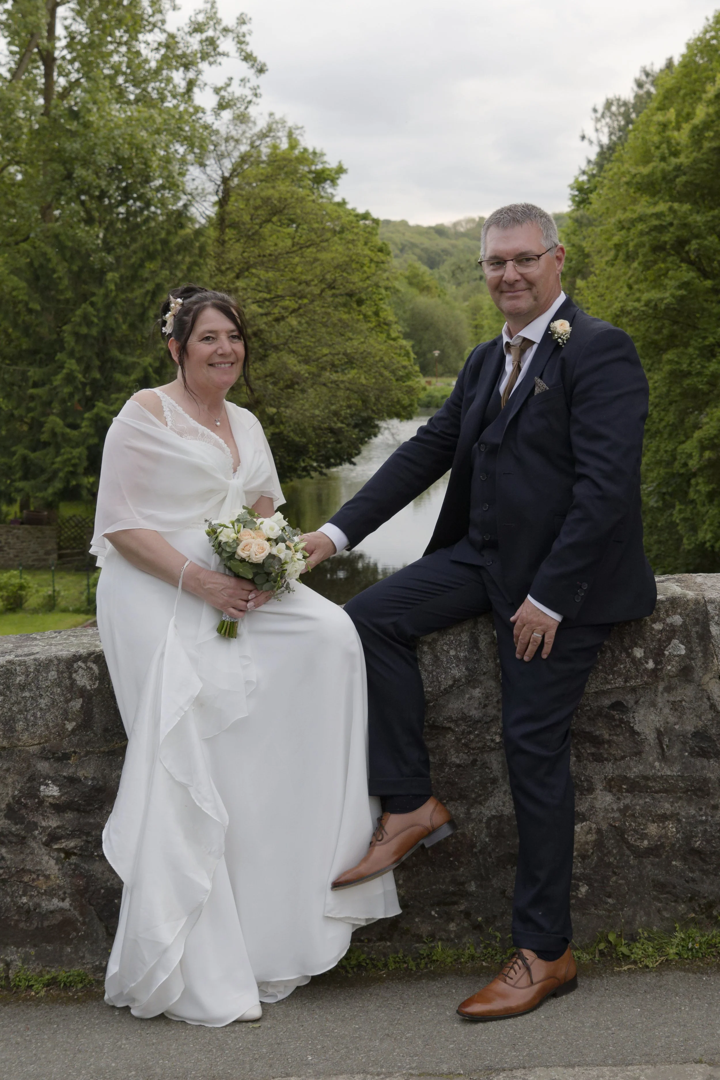 Un couple lors d'un mariage, la femme en robe blanche et le homme en costume sombre, se tenant la main sur un mur en Pierre, entourés de nature verte.