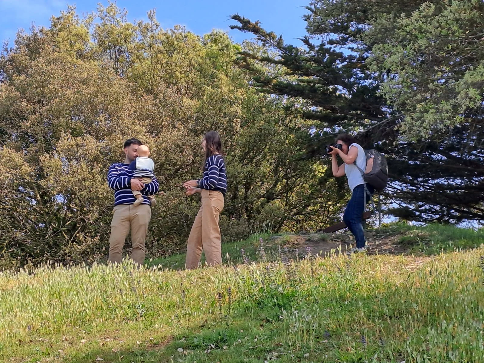 Une famille en pleine séance photo dans un parc avec des arbres en arrière-plan. Un homme tient un bébé, une femme parle avec lui, et une autre personne prend la photo.