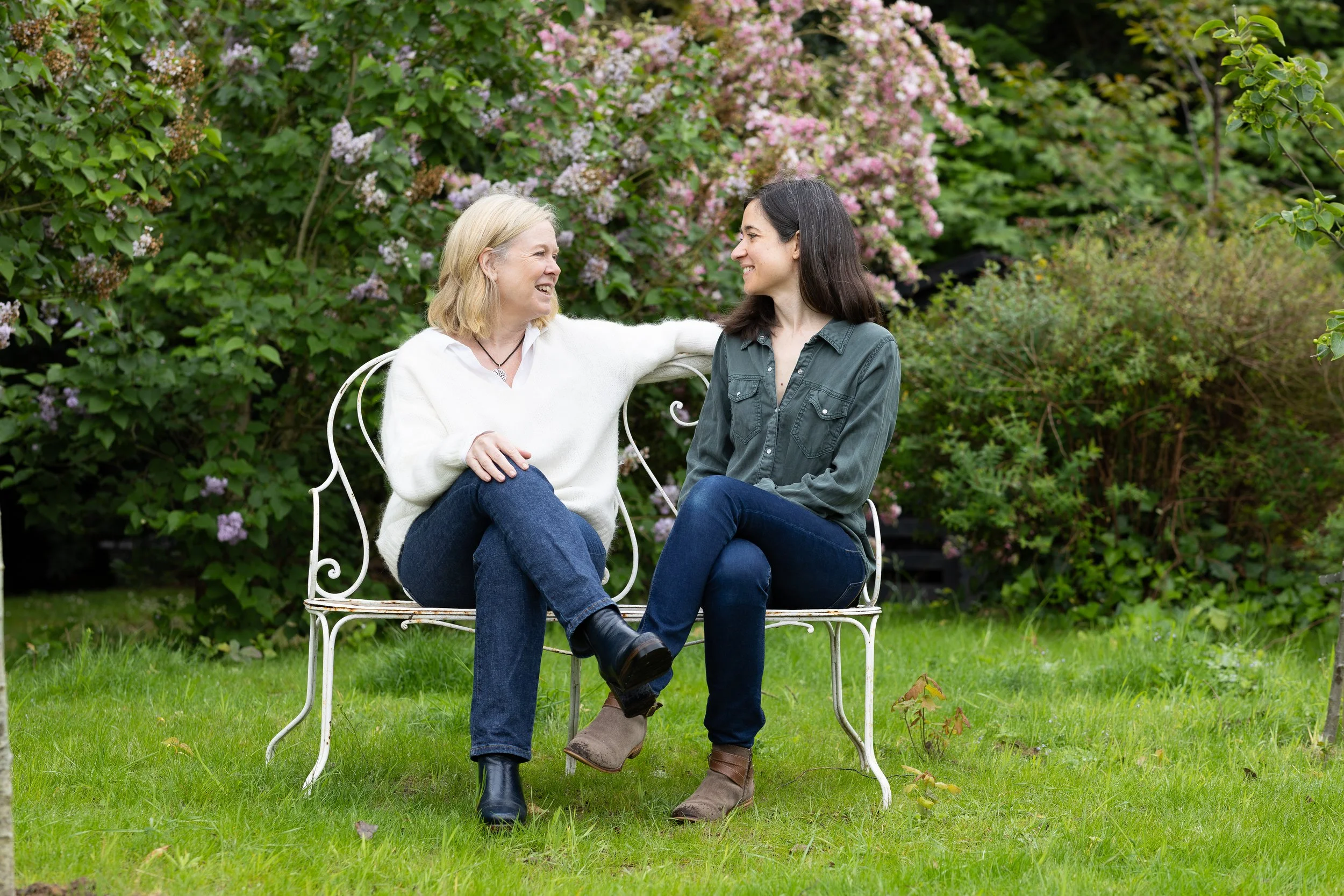 Deux femmes assises sur un banc en métal dans un jardin avec des buissons en fleurs roses et blanches. Elles se regardent et sourient, l'une a les cheveux blonds et l'autre a les cheveux bruns.