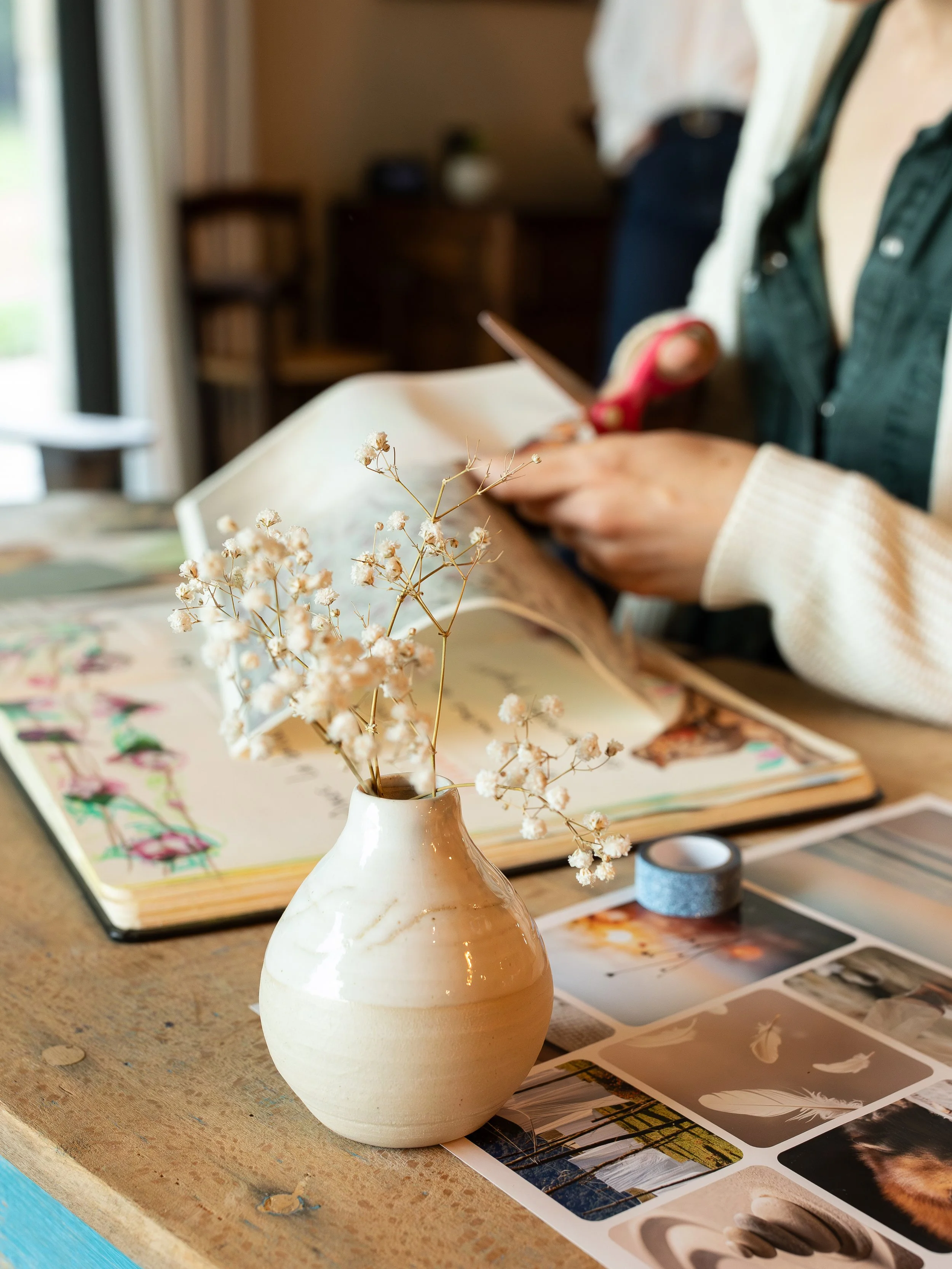 Un vase beige avec des fleurs séchées blanches placé sur une table avec des magazines et des échantillons de peinture. En arrière-plan, une personne regarde un téléphone portable.