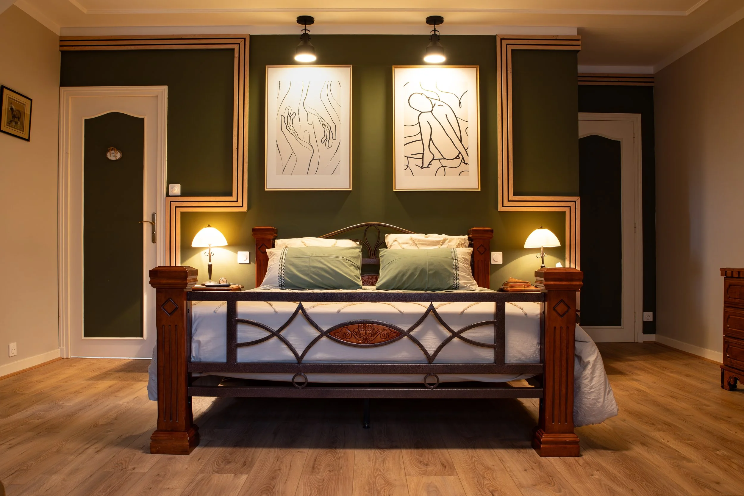 Chambre avec lit en bois, deux lampes de chevet, deux tableaux abstraits au-dessus de la tête de lit, murs verts et beige, sol en bois.