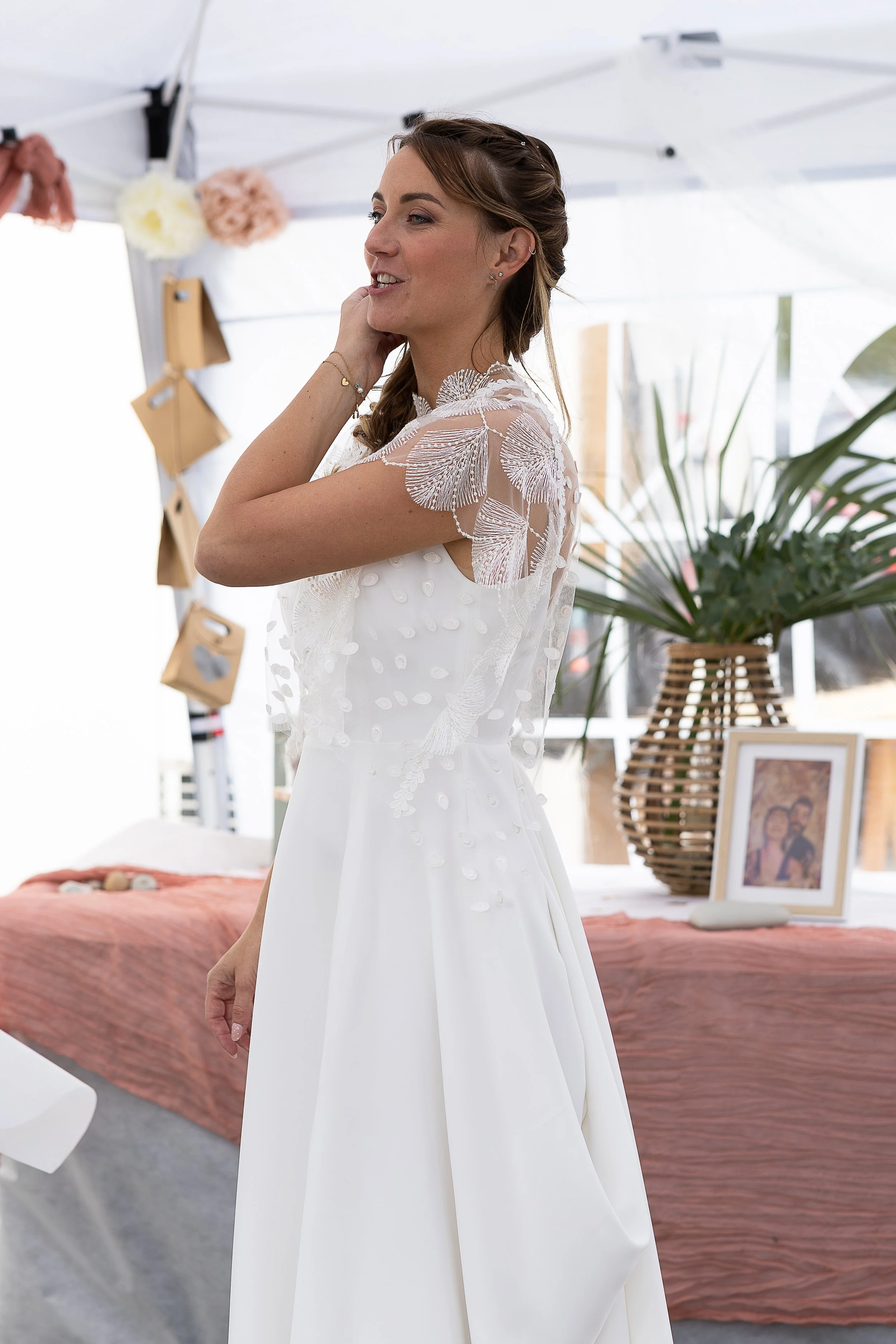 Femme blonde en tenue de mariée blanche se caresse les cheveux. Elle est debout sous un chapiteau décoré pour l'occasion.