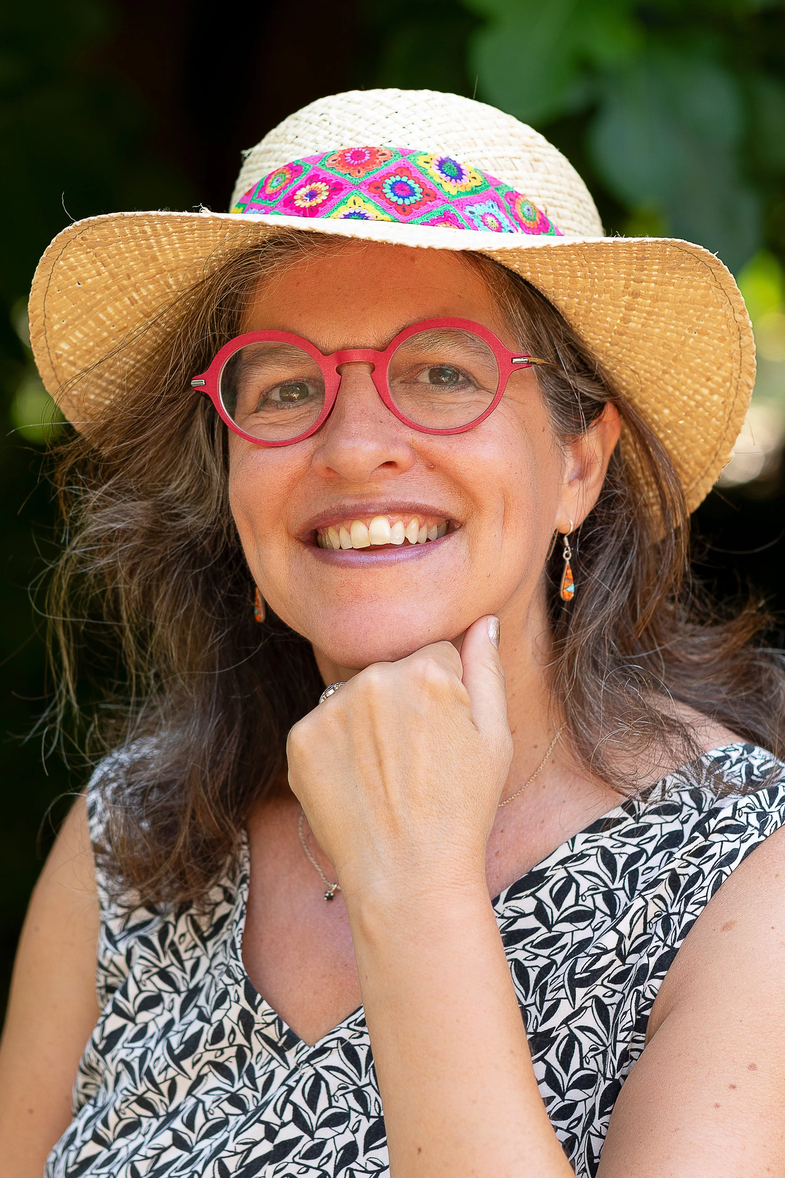 Femme souriante portant un chapeau de paille, lunettes roses, et une veste à motif floral, en extérieur.