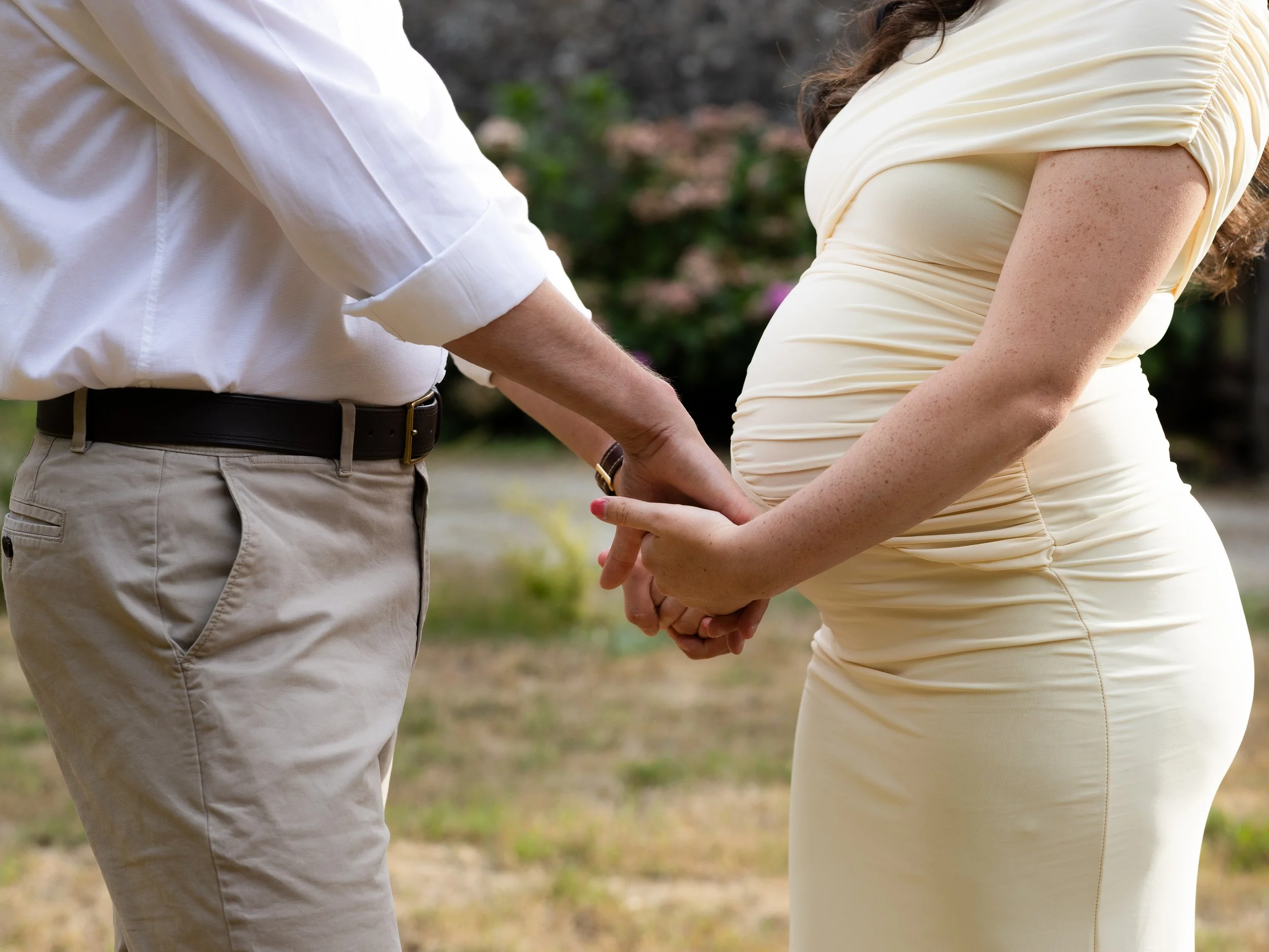 Un couple se tenant la main dans un parc. On ne voit pas les visages, la femme enceinte porte une robe beige moulante et l'homme habillé en blanc porte un pantalon beige.