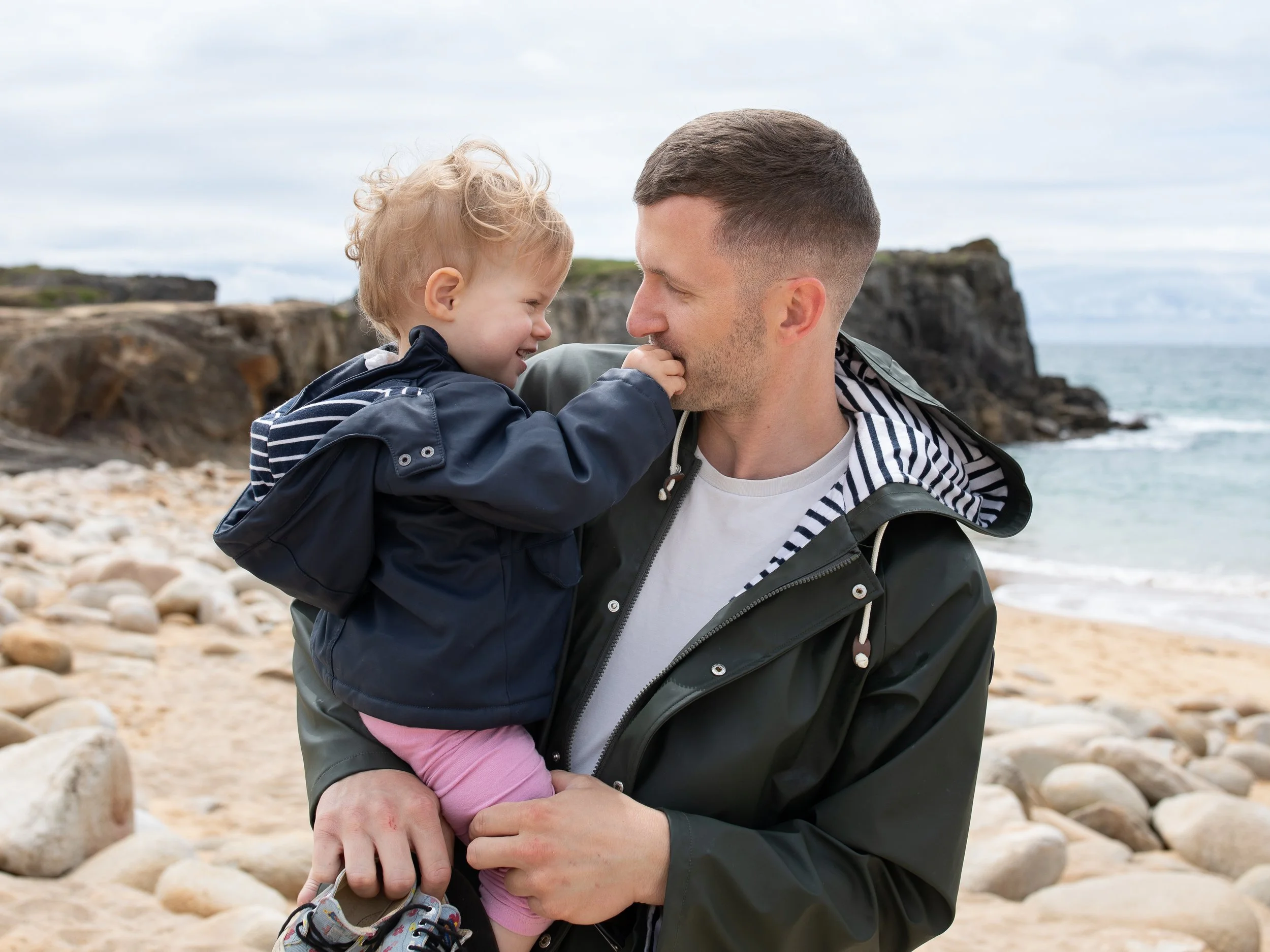 Un père et sa petite fille dans les bras se regardent  sur la plage avec des rochers et la mer en arrière-plan.