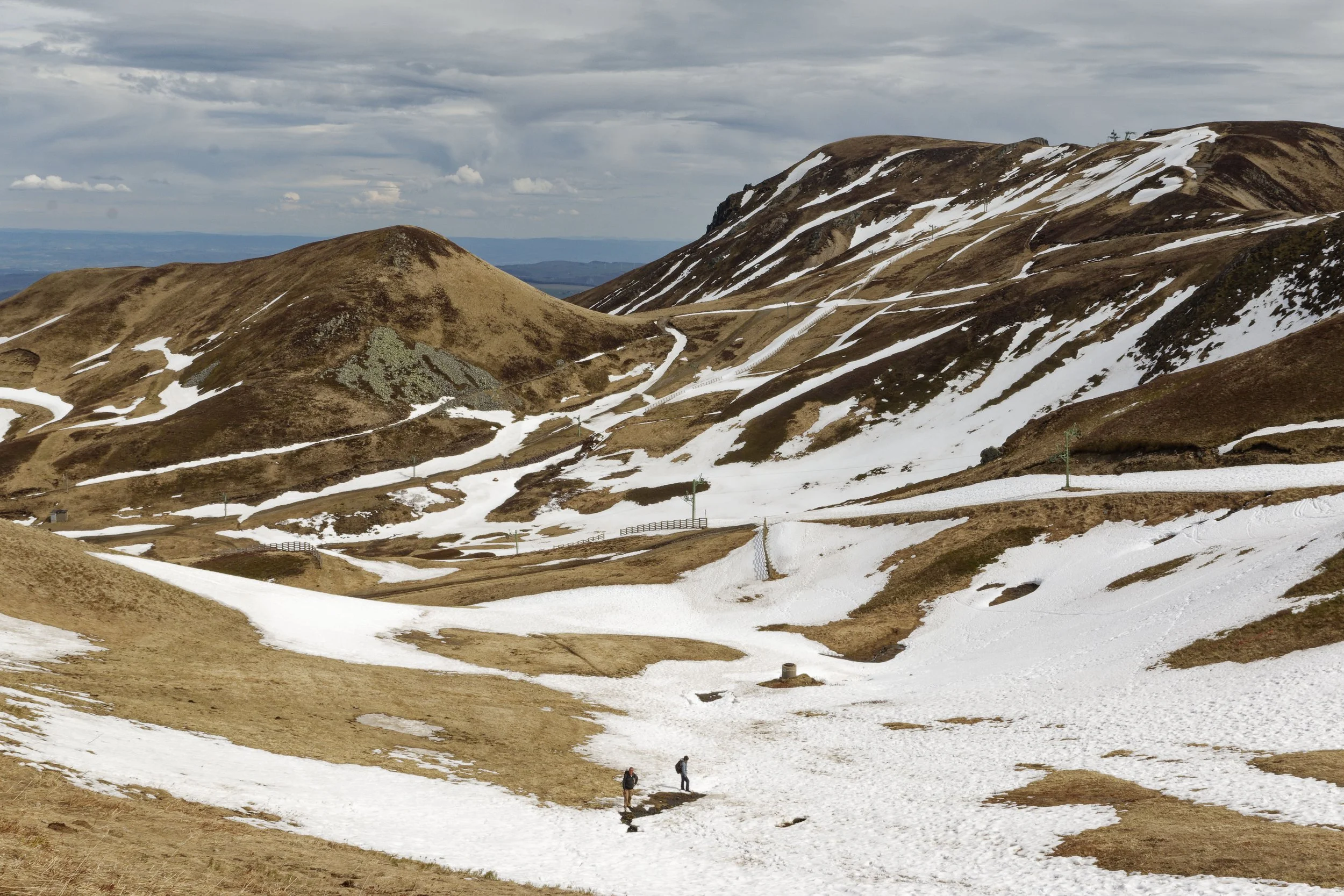 Photo de paysage de montagne, la neige est présente sur les routes, la terre est jaune. On aperçoit des marcheurs au loin.
